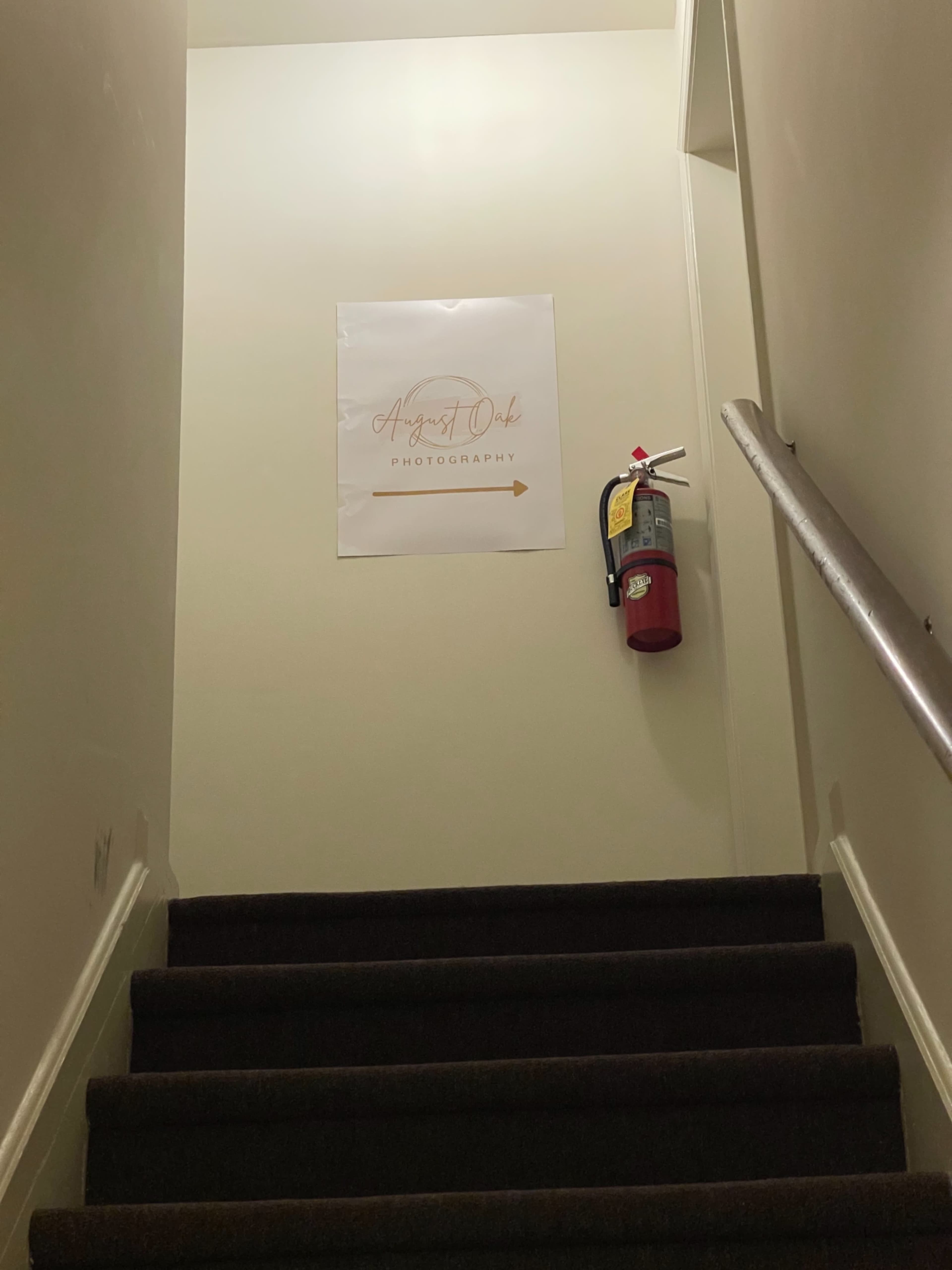 A fire extinguisher is mounted on a wall next to a sign directing to "August Dark Photography" at the top of a staircase.