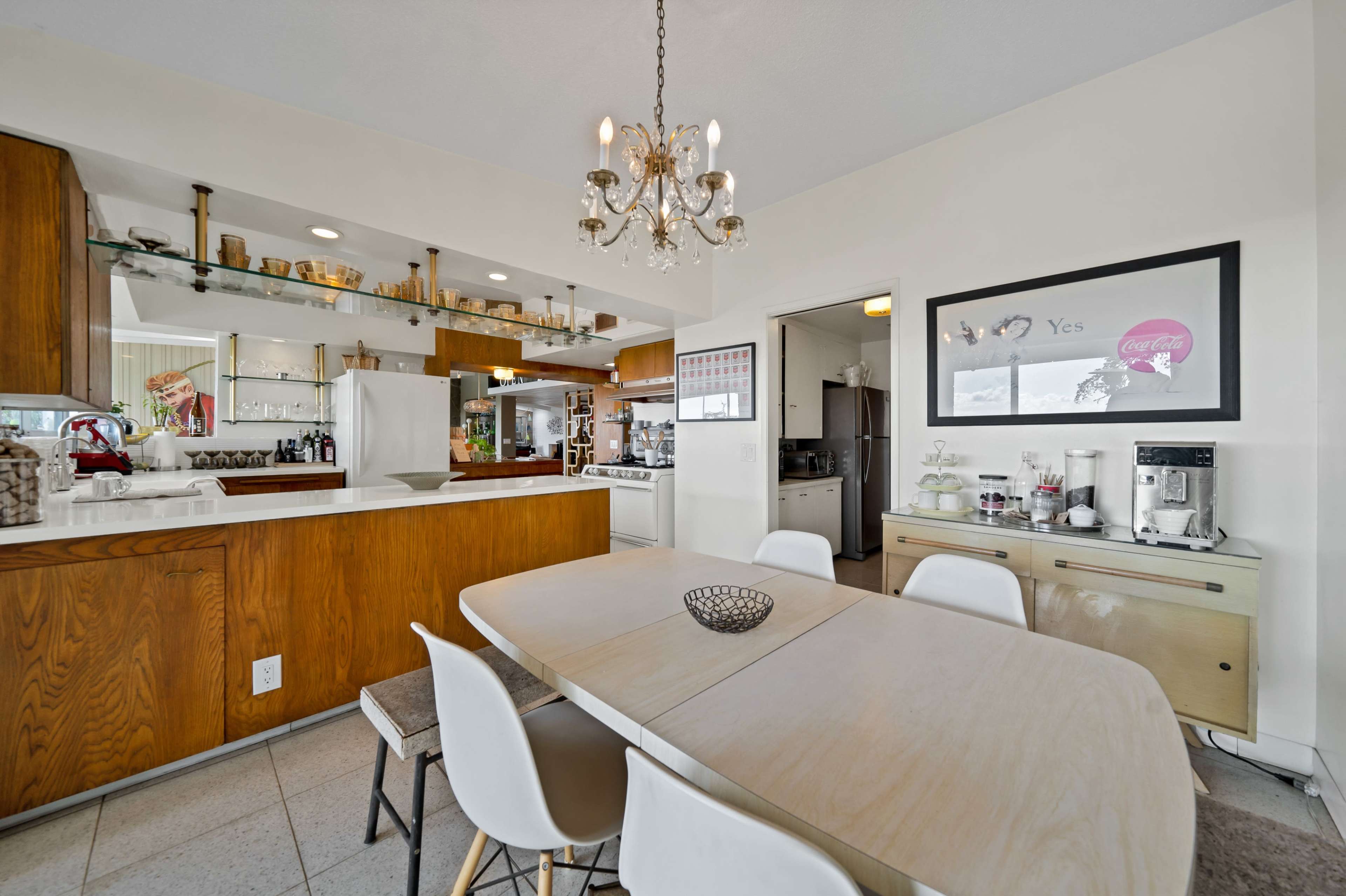 The image shows a modern kitchen with a central dining table surrounded by light-colored chairs and wooden cabinetry, featuring various kitchen appliances and decor items.