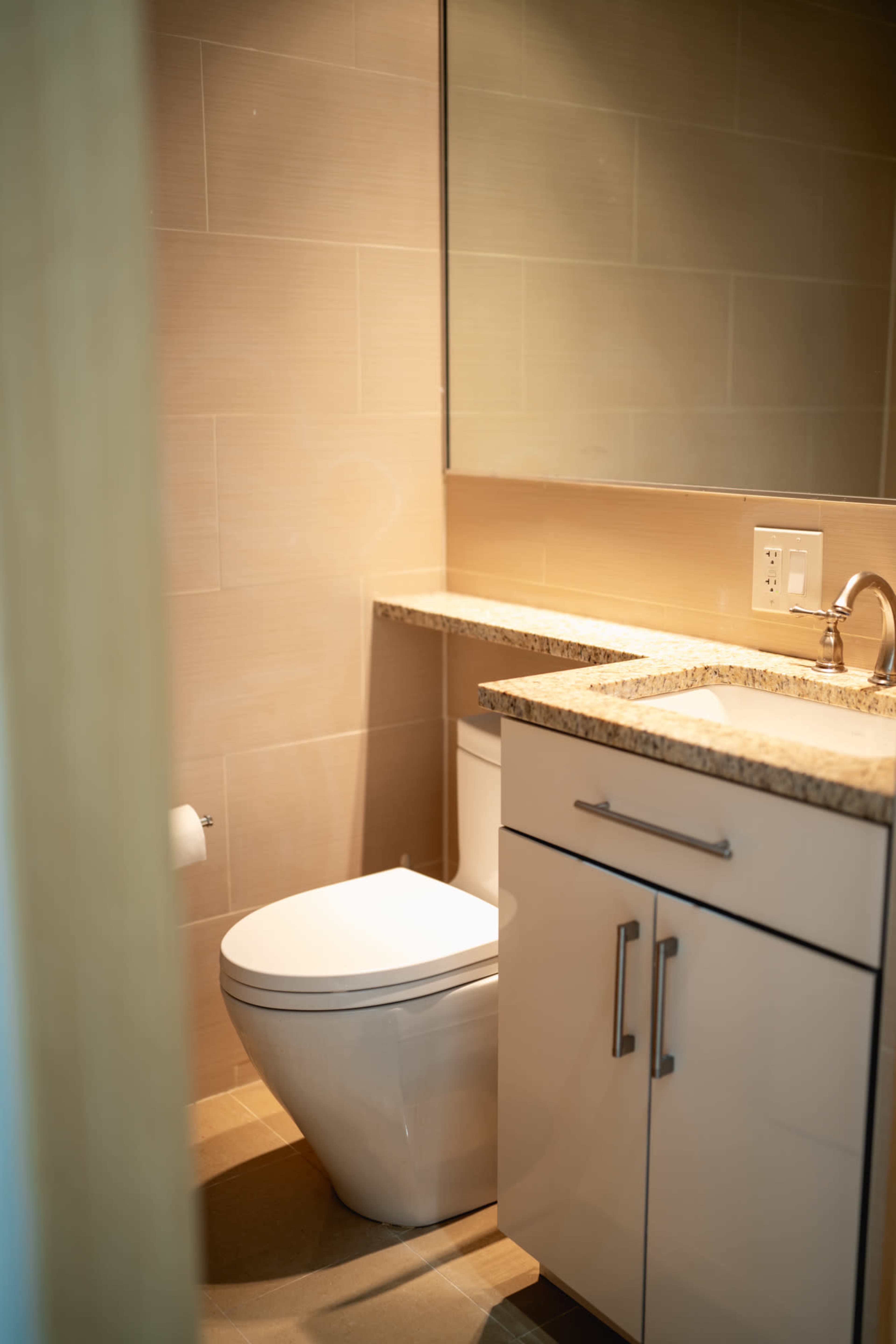 The image shows a modern bathroom featuring a toilet, a sink with a granite countertop, and a large mirror on the wall.