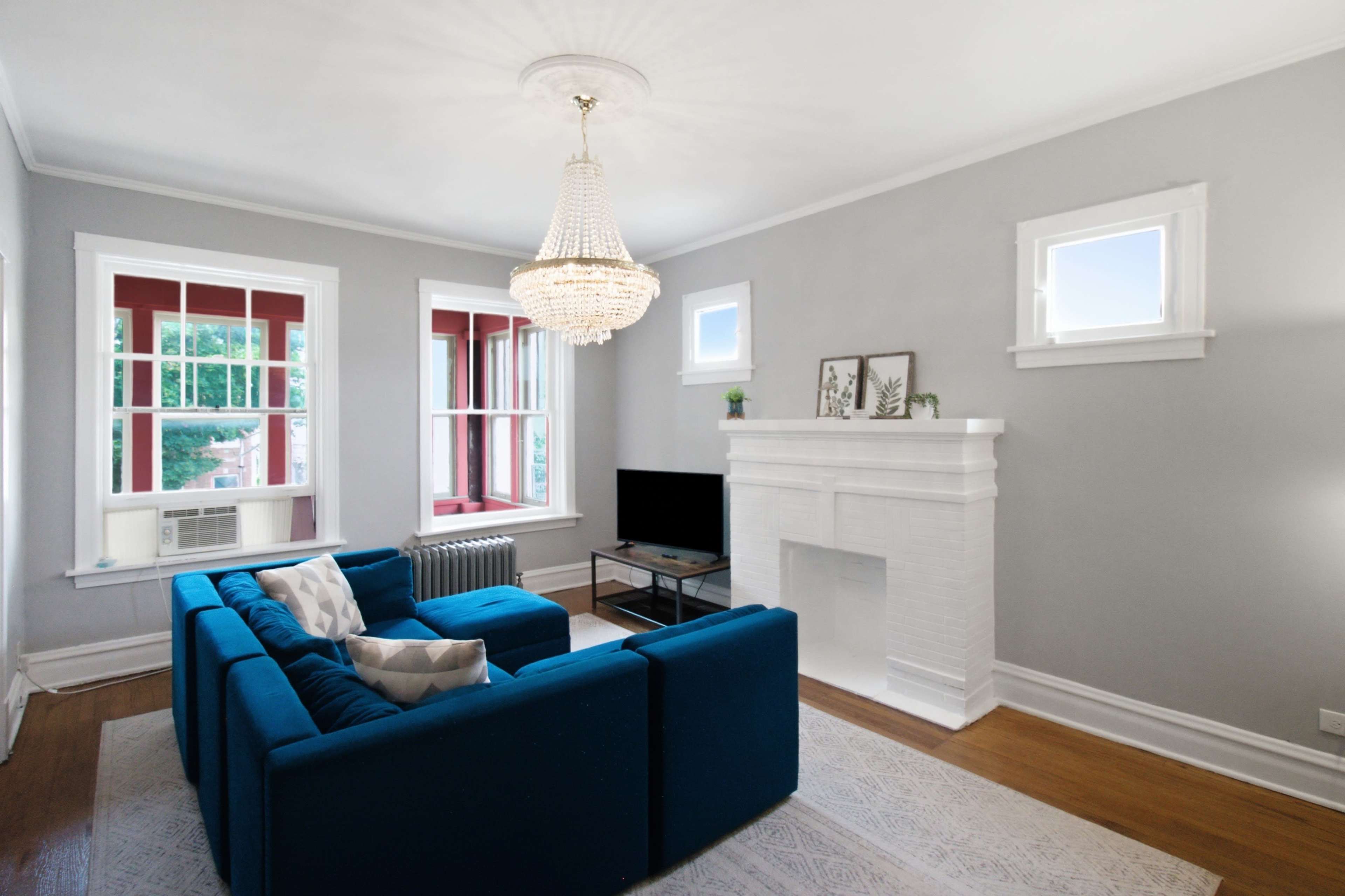 The image shows a living room featuring a blue sofa, a television on a black stand, a white fireplace, and large windows with red trim.