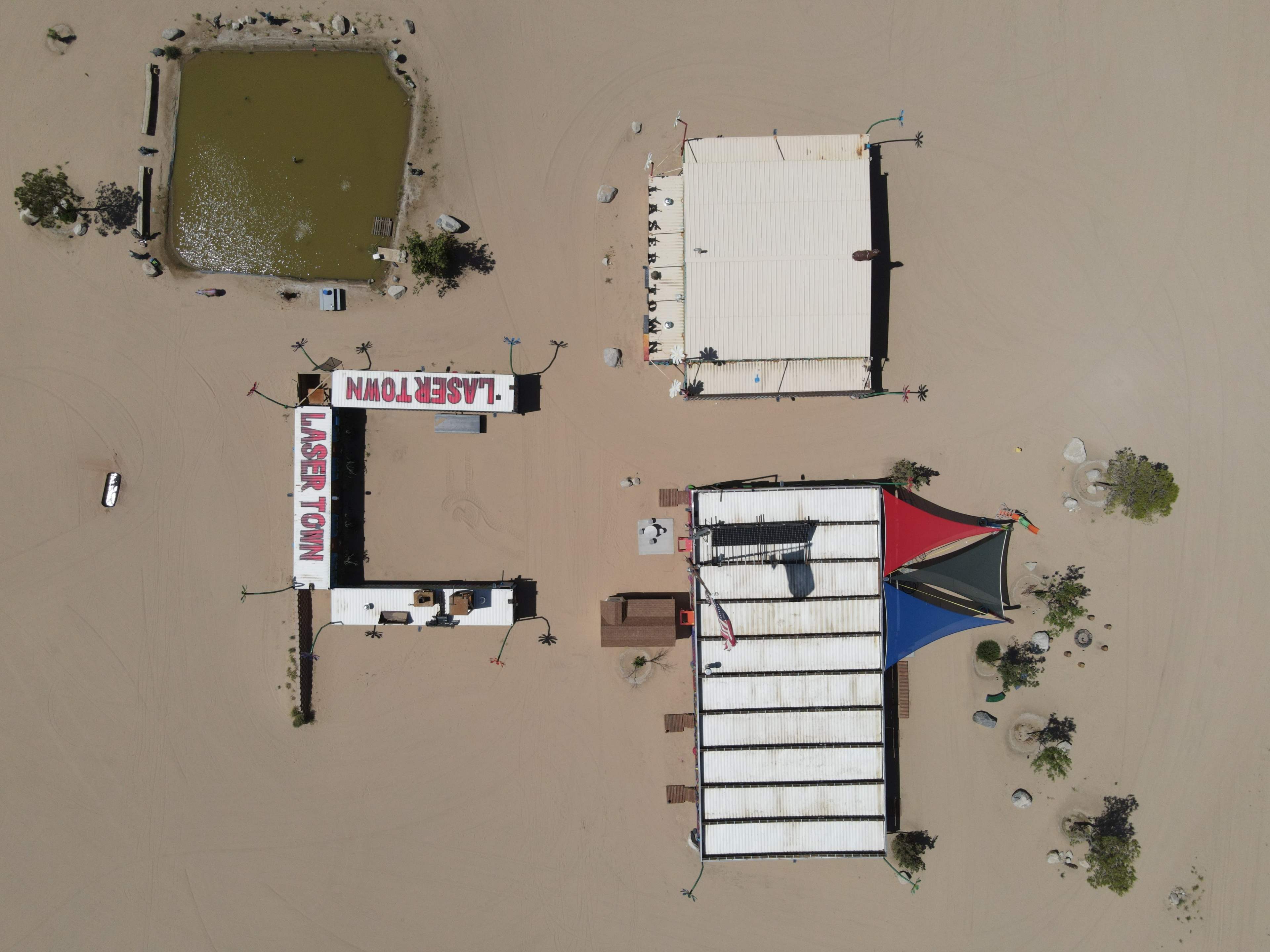 An aerial view of a desert landscape featuring several structures, including a building labeled "LASERTOWN," a pond, and a multi-roofed building.