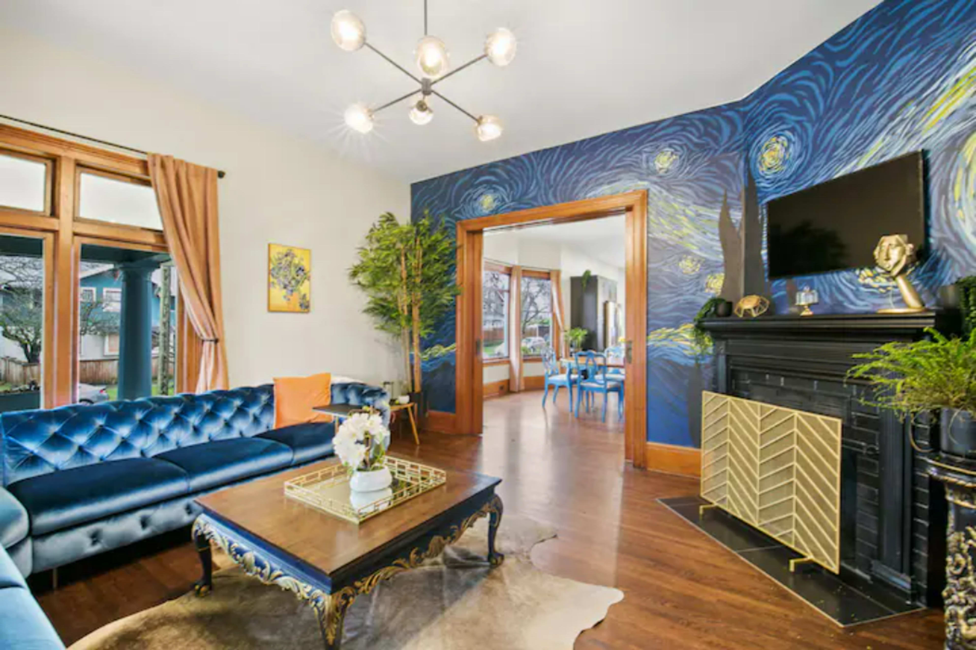 A living room features a blue couch, a coffee table, and a black fireplace, with a vibrant wall mural and a dining area visible through an open doorway.