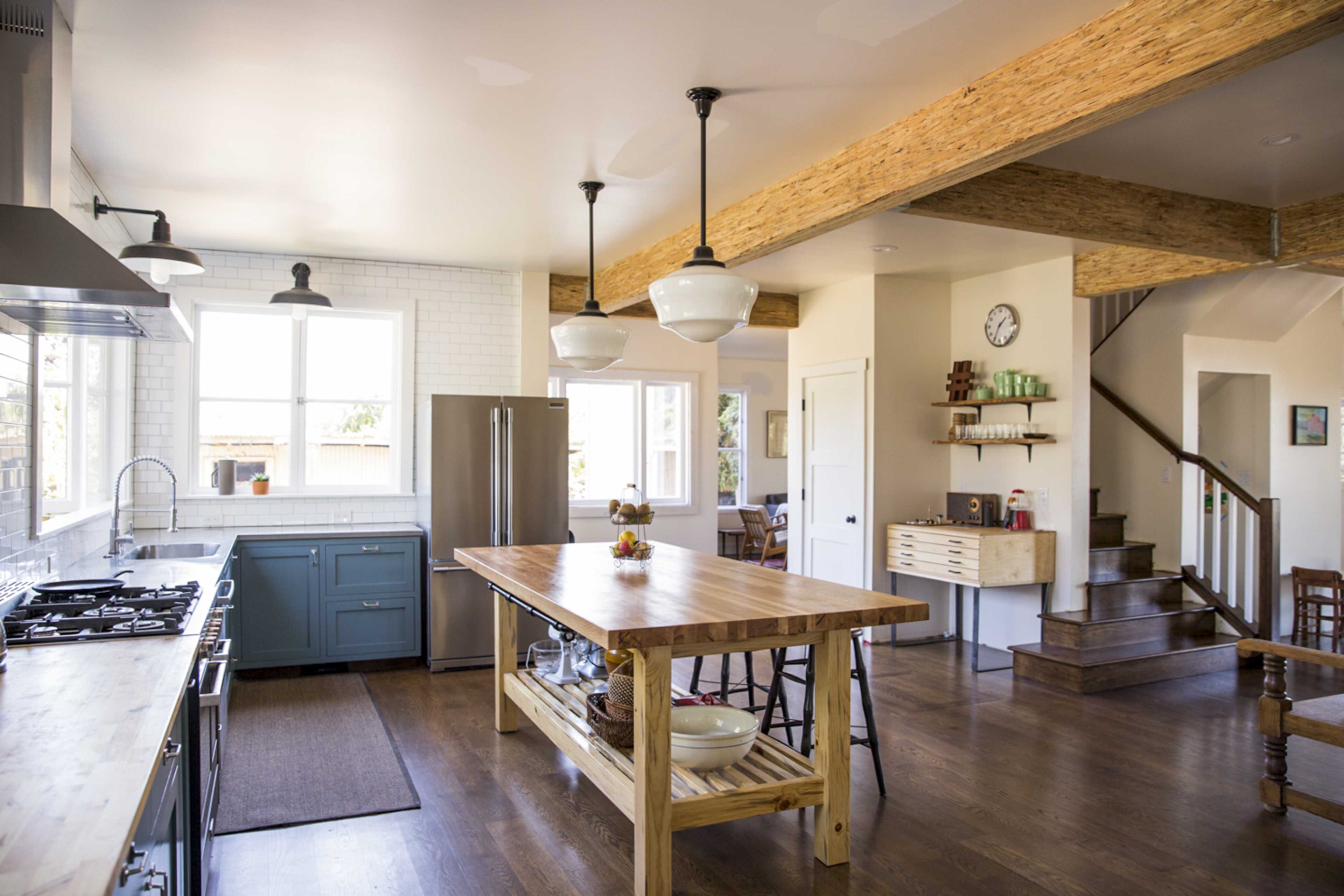 The kitchen features a large center island with a wooden countertop, blue cabinets, stainless steel appliances, and open shelving, alongside a staircase leading to another level.