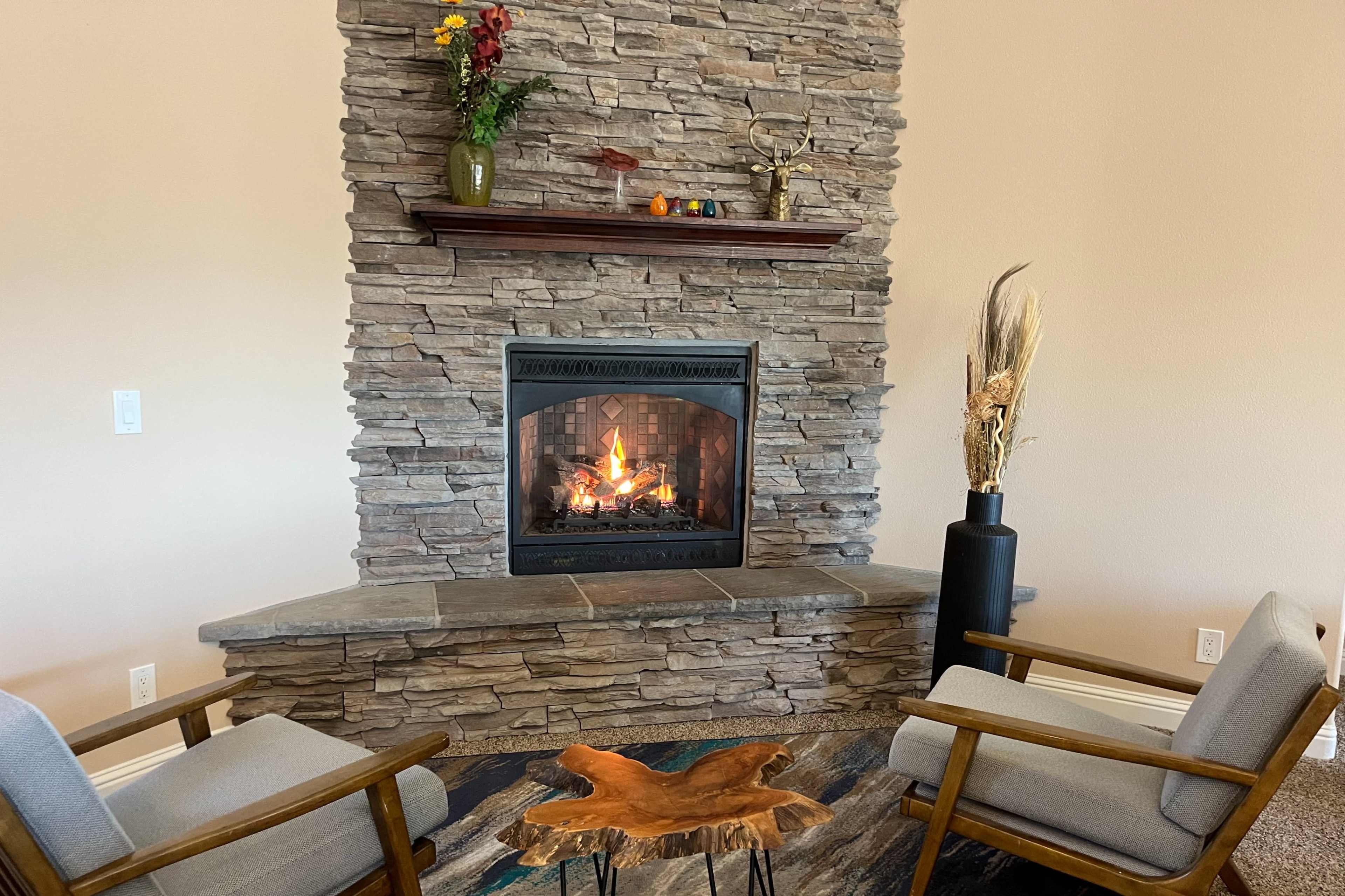 A stone fireplace with a roaring fire is flanked by two chairs and a small wooden table, set against a beige wall.