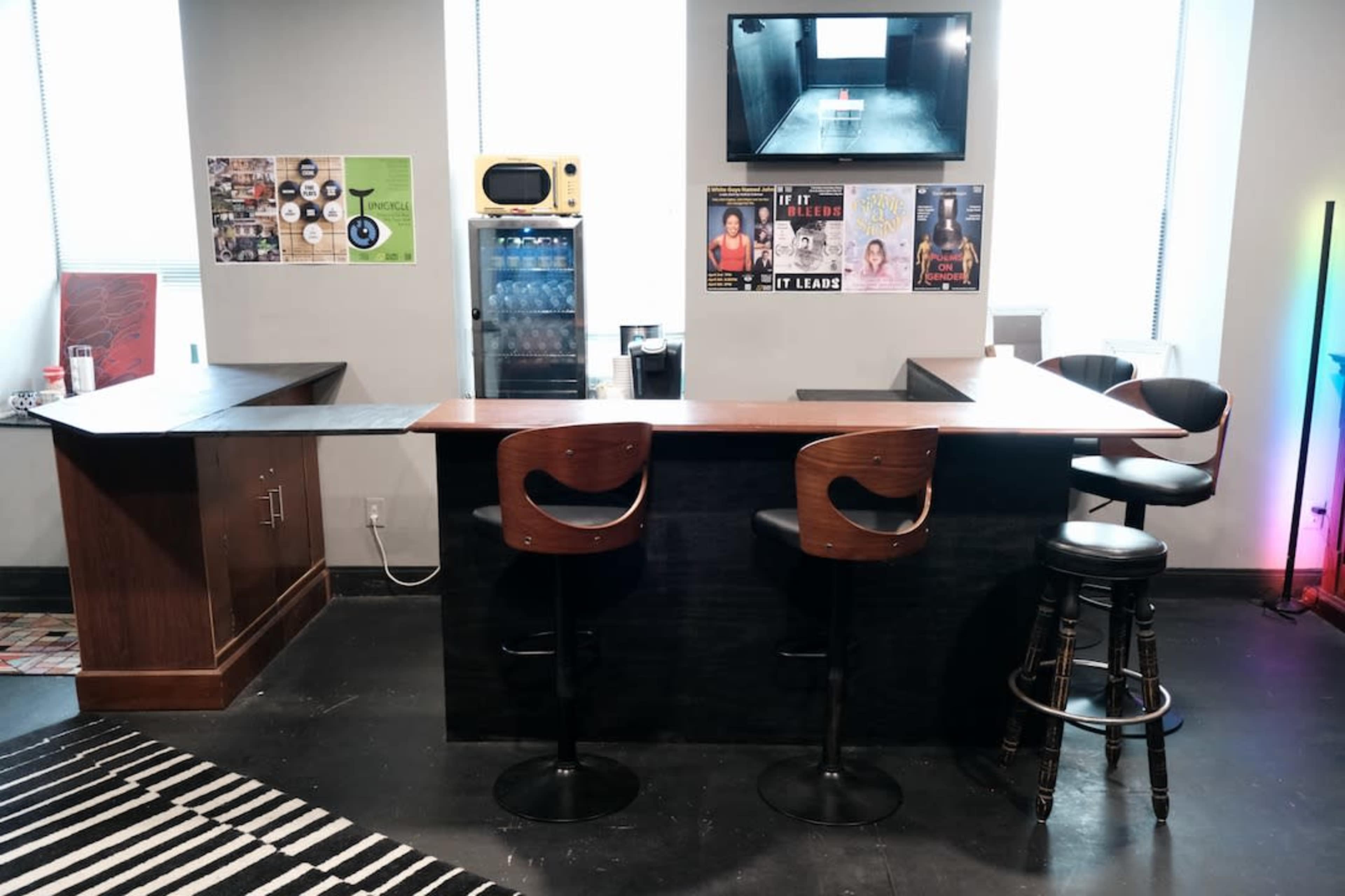 The image shows a modern bar area with a wooden countertop, three chairs, a retro-style television screen mounted on the wall, and various posters and decorations around the space.