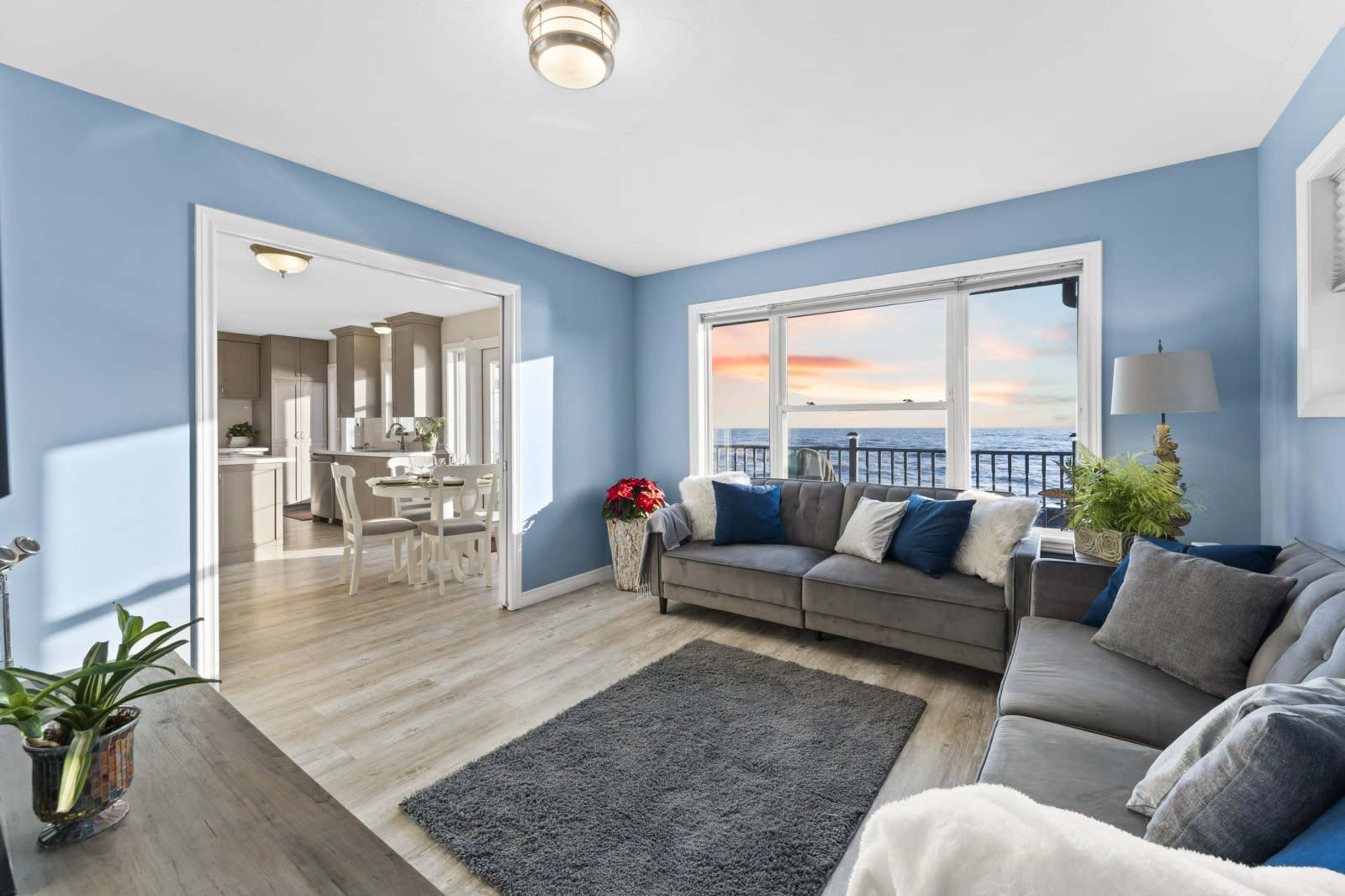 A living room with blue walls, a gray sofa, and large windows that overlook a sunset by the water.