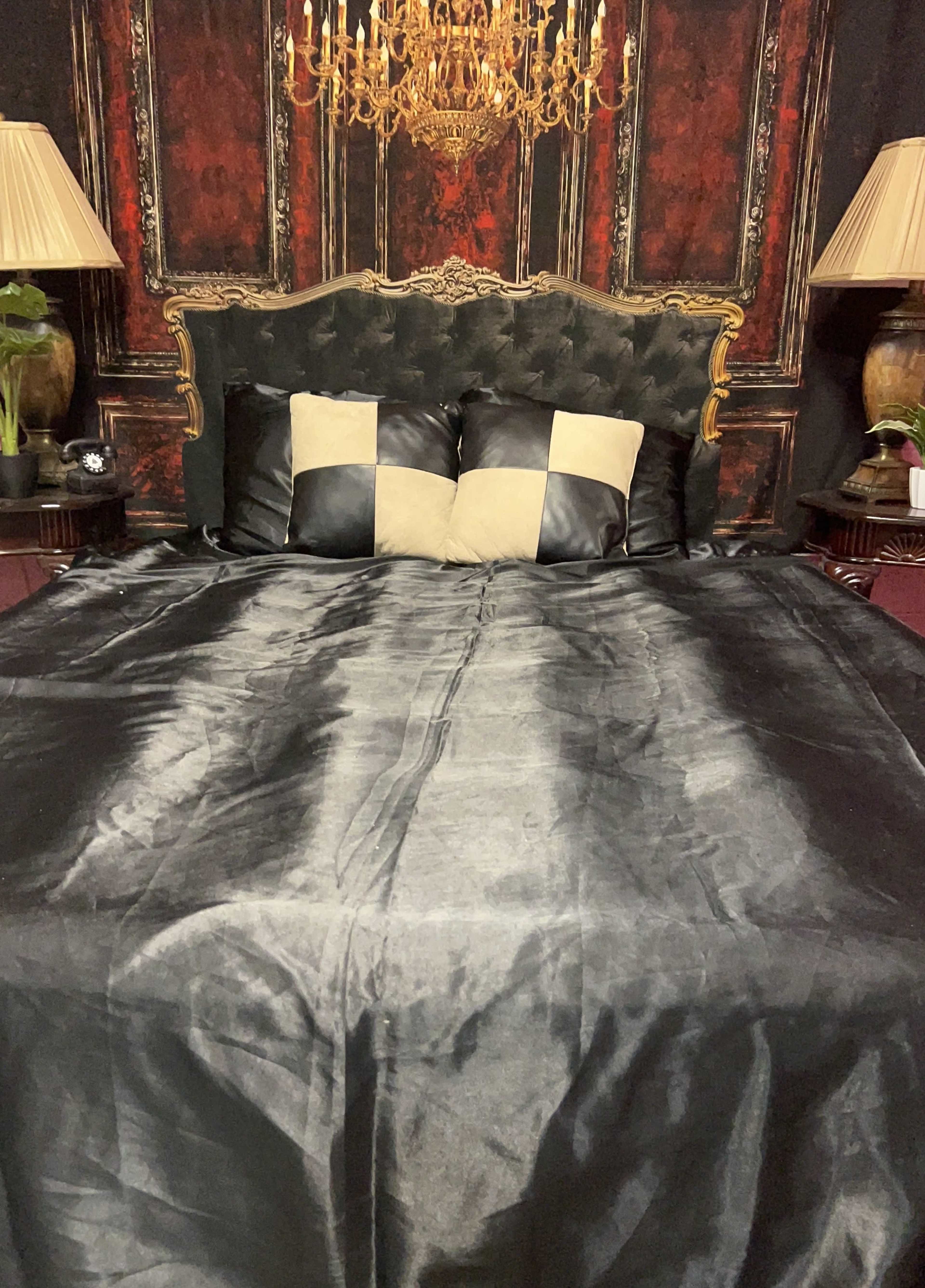 The image shows a styled bed with a dark satin cover and two square pillows in a black and beige checkerboard pattern, set against a richly decorated wall and accompanied by ornate lamps.