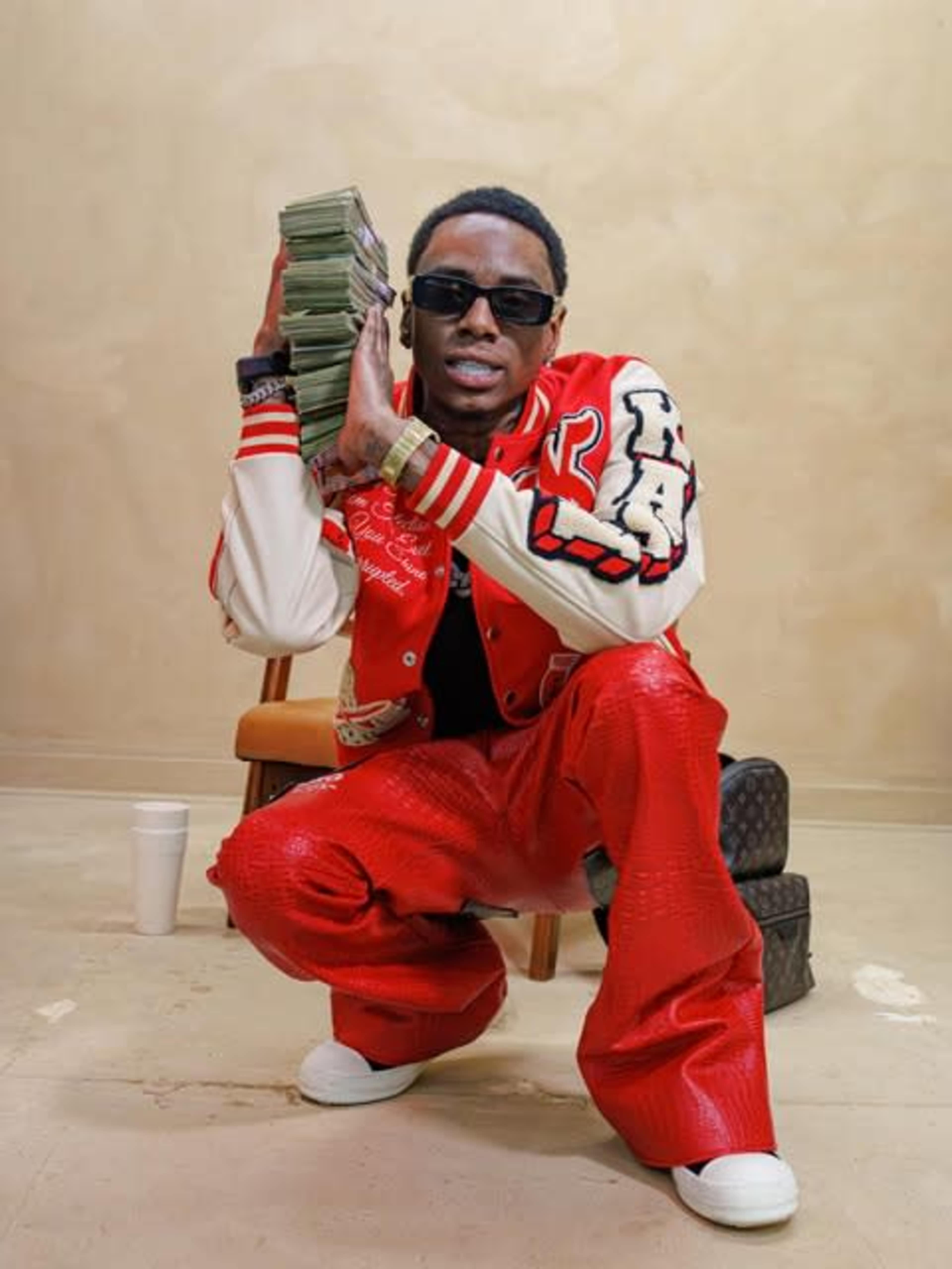 A person dressed in a red outfit and sunglasses poses while holding a stack of cash in a minimalistic indoor setting.