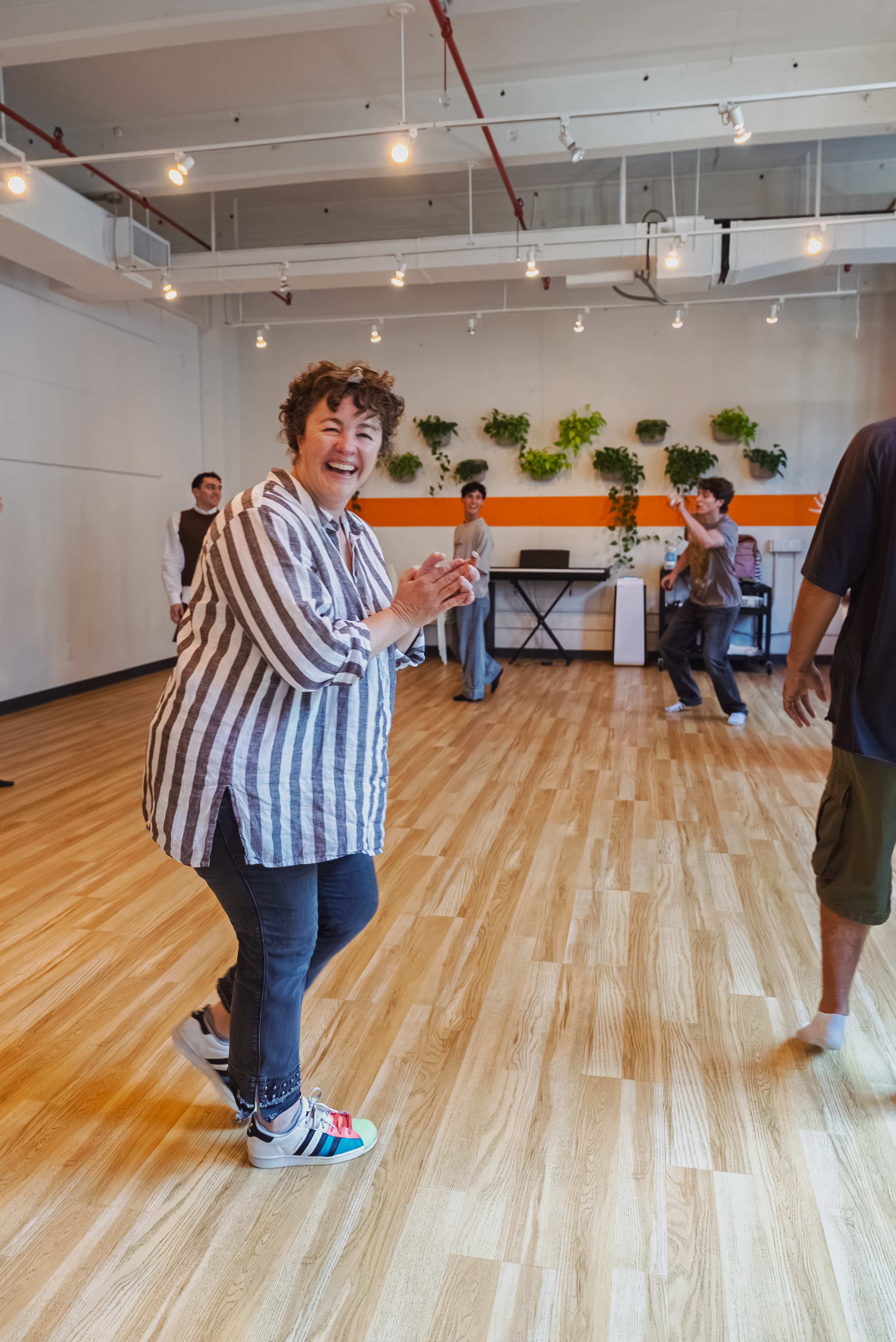 A group of people are dancing in a spacious studio with wooden floors and green plants on the walls, while one woman with curly hair smiles and claps her hands.