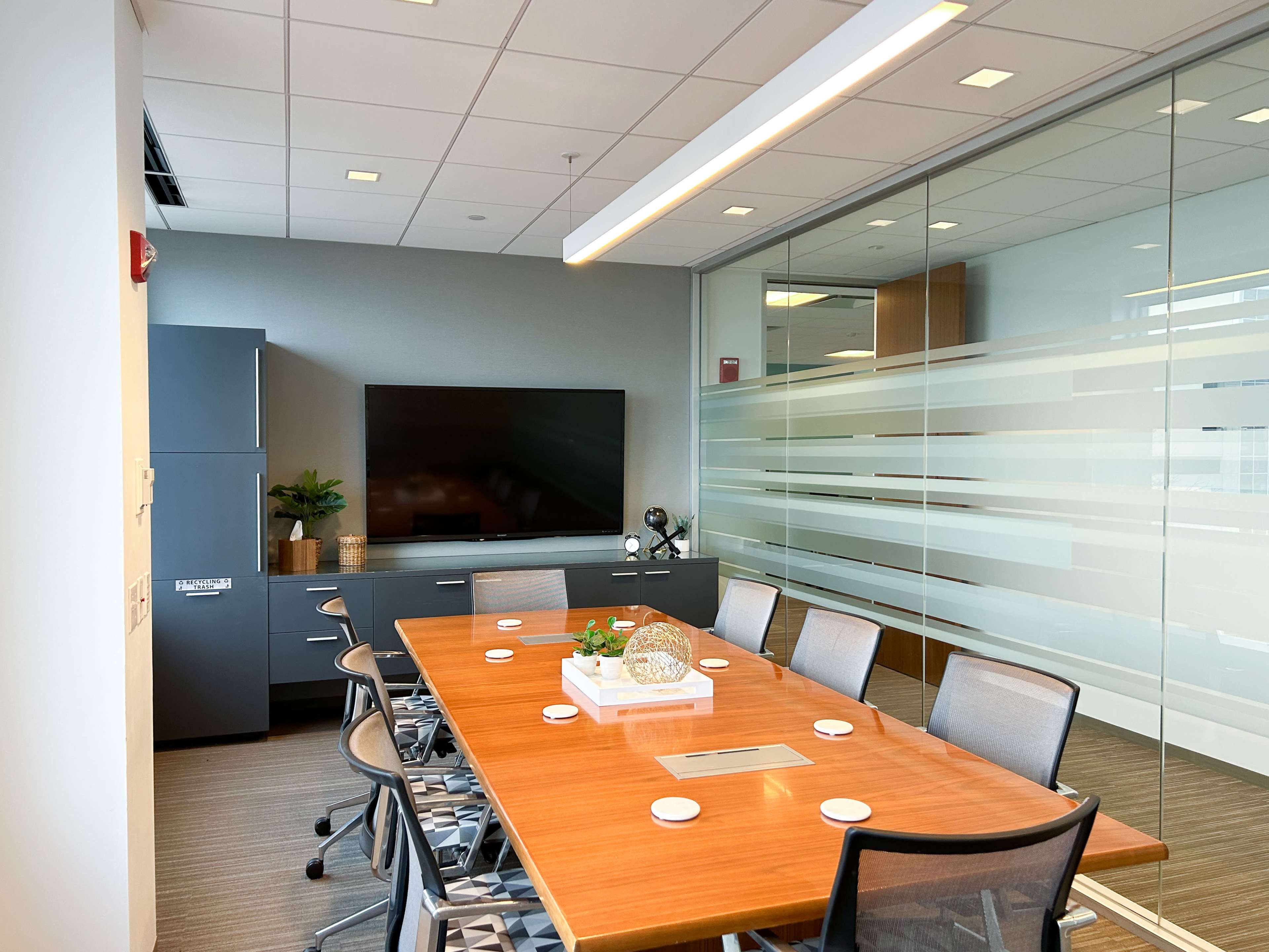 A modern conference room features a large wooden table surrounded by chairs, with a wall-mounted TV and glass partitions providing light.