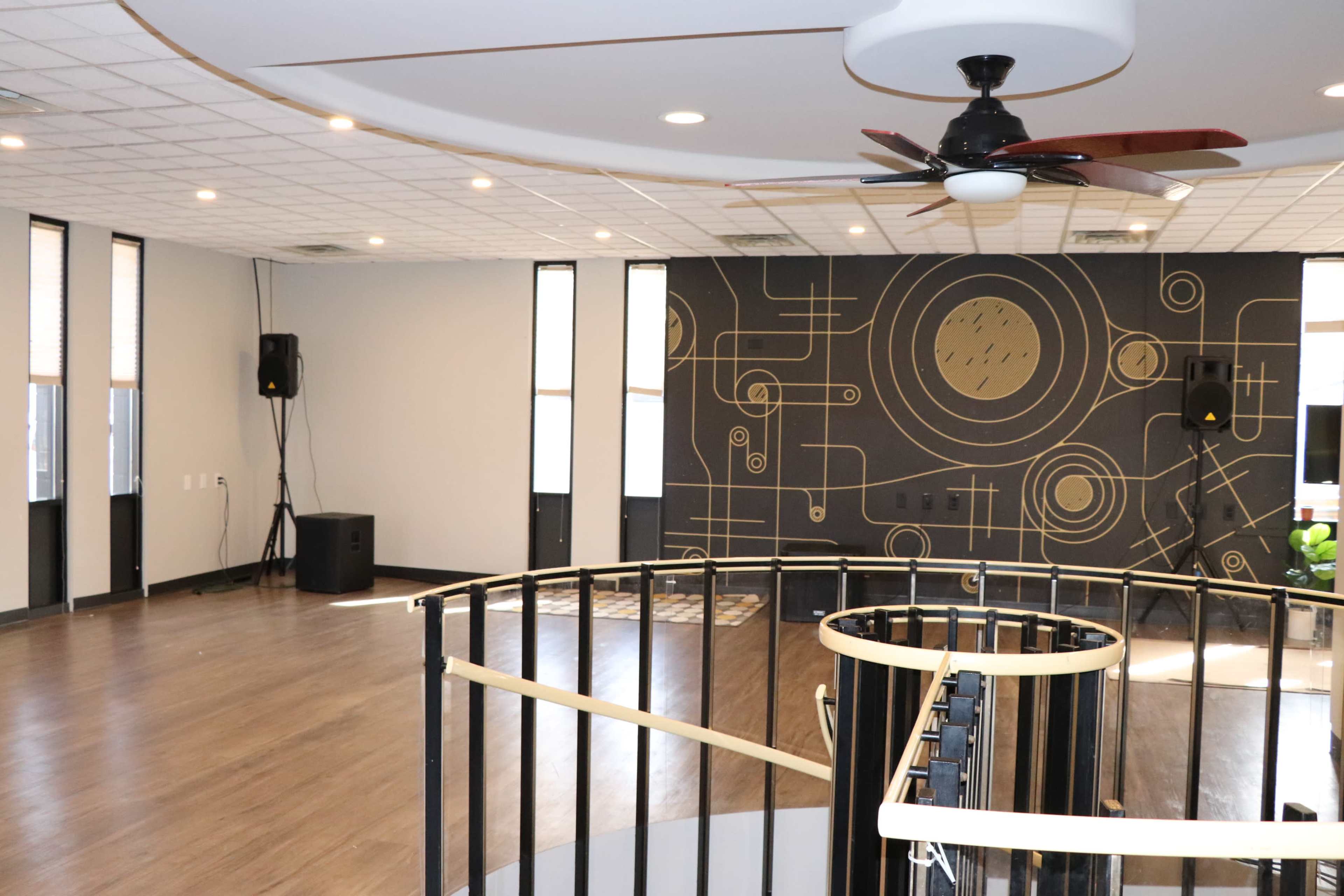 The image shows an empty room with a curved railing, a ceiling fan, a dark wall featuring geometric designs, and large windows allowing natural light.