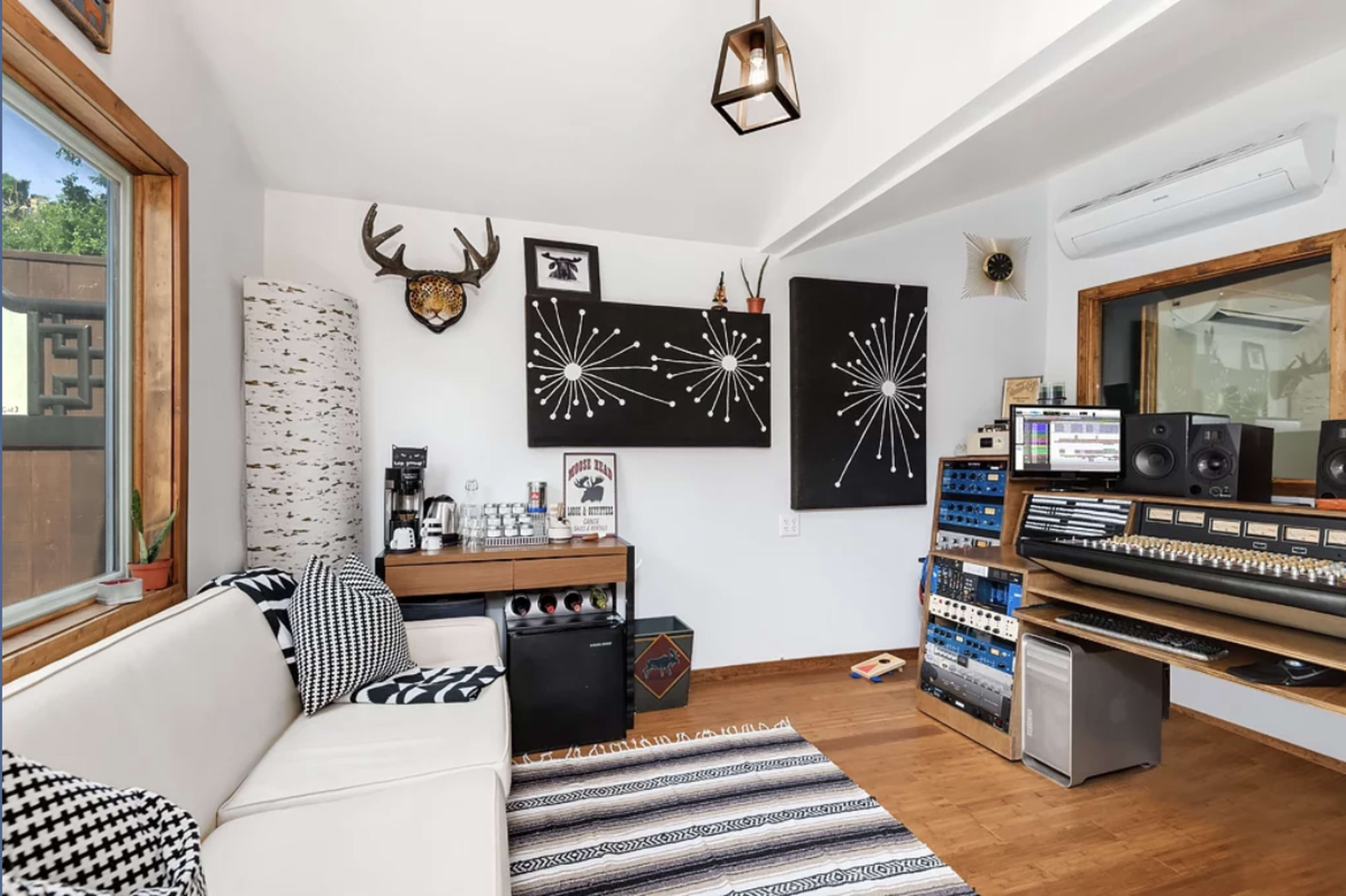The image depicts a modern, minimalist studio space featuring a white couch, a wooden desk with a coffee maker, and audio equipment against a backdrop of black and white wall art.