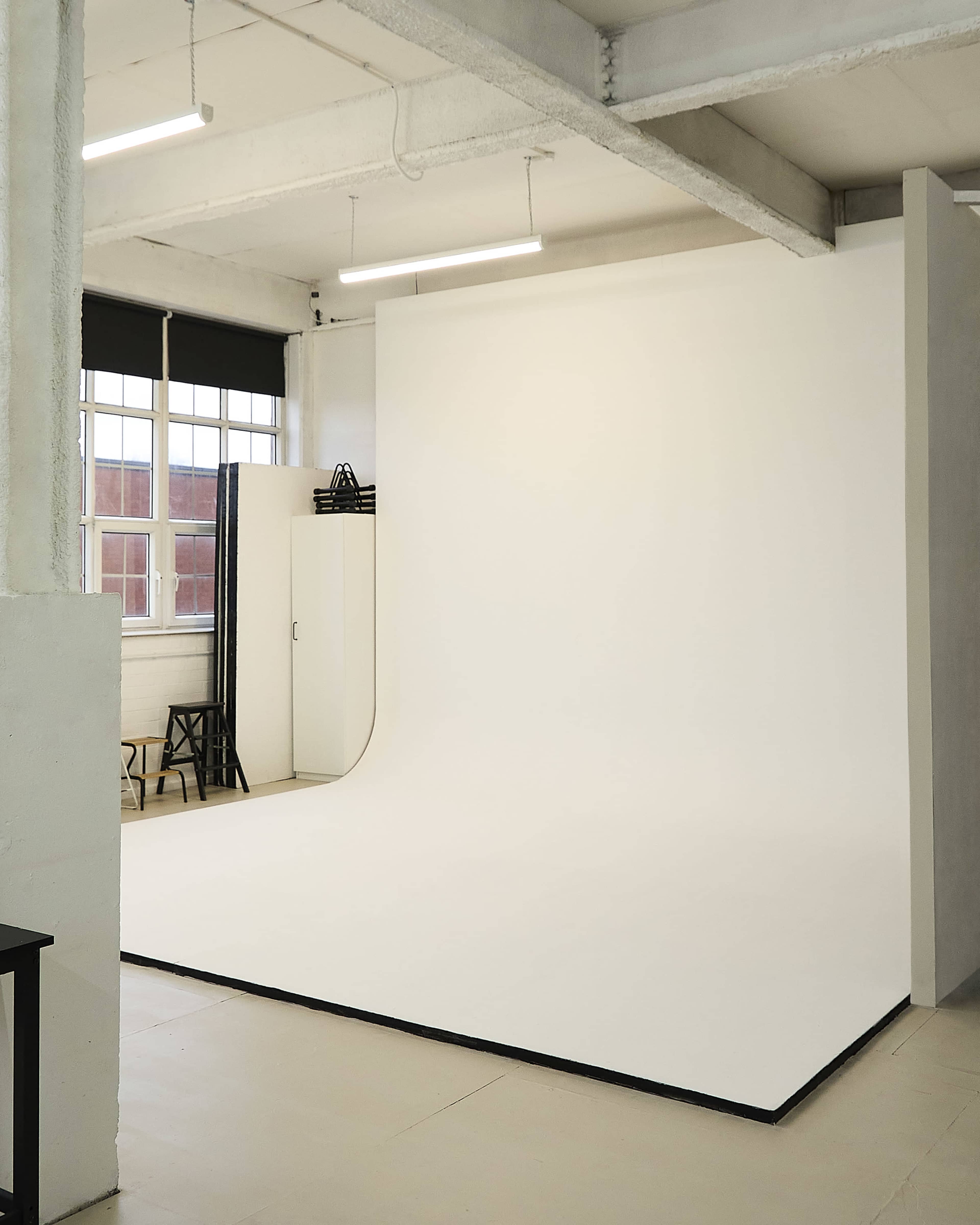 The image shows a spacious photography studio featuring a large white backdrop, a cabinet, and a black chair against a well-lit interior.