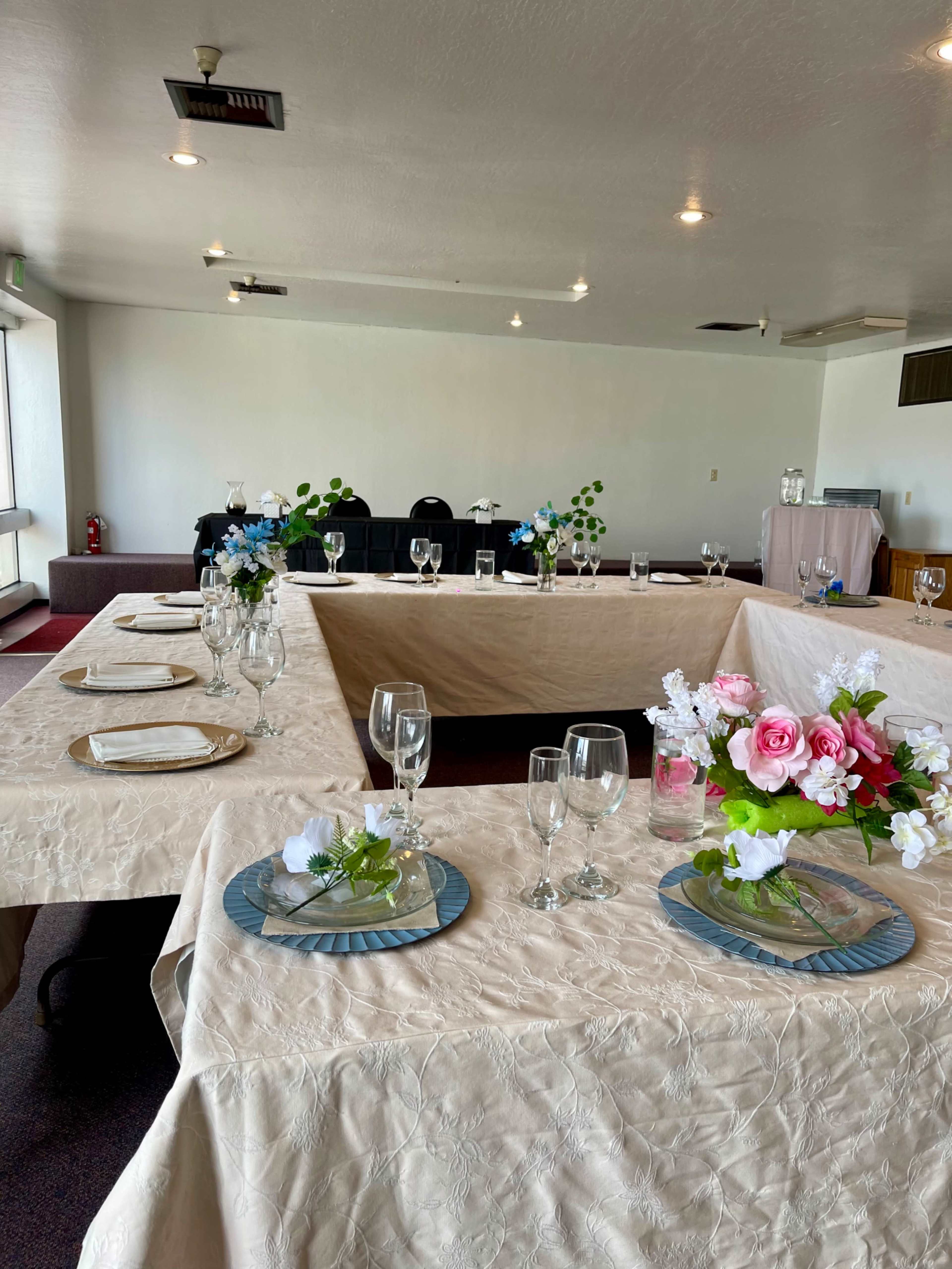 A dining area set with tables covered in light-colored tablecloths, each adorned with centerpieces of flowers and glassware.