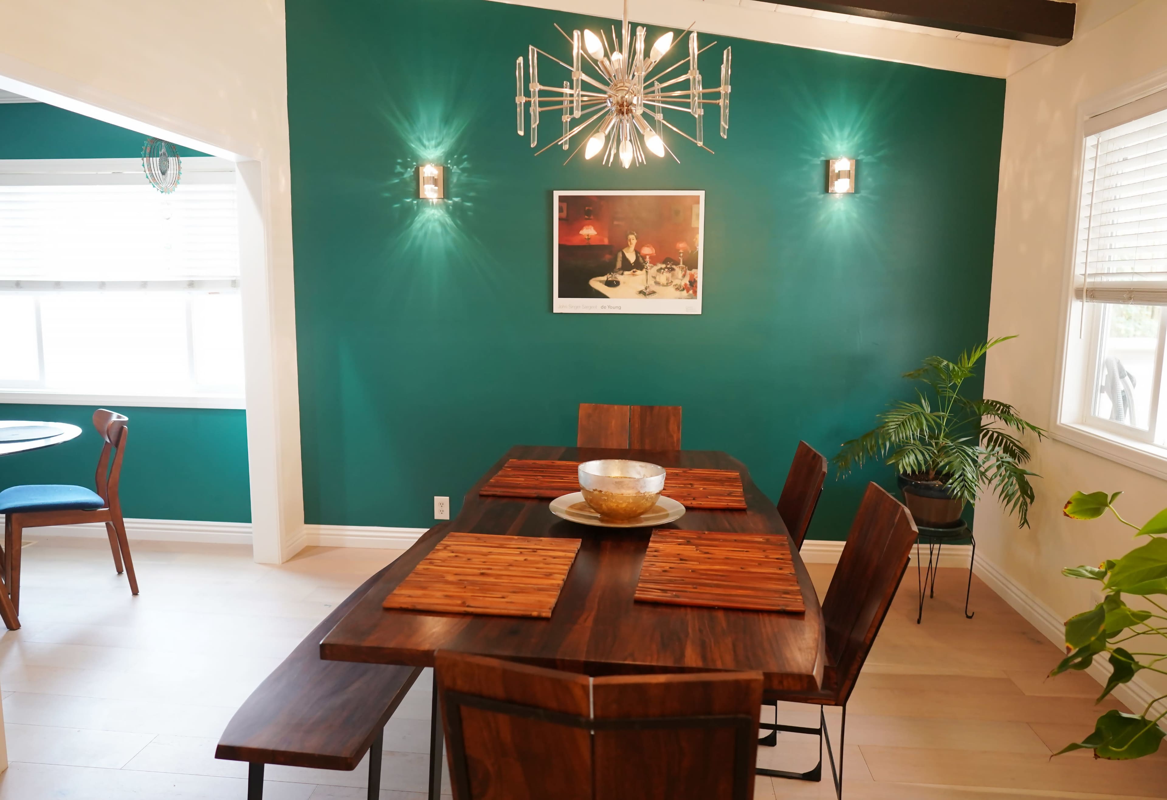 A dining area features a wooden table with placemats, surrounded by chairs, against a teal wall with a chandelier and a framed photograph.