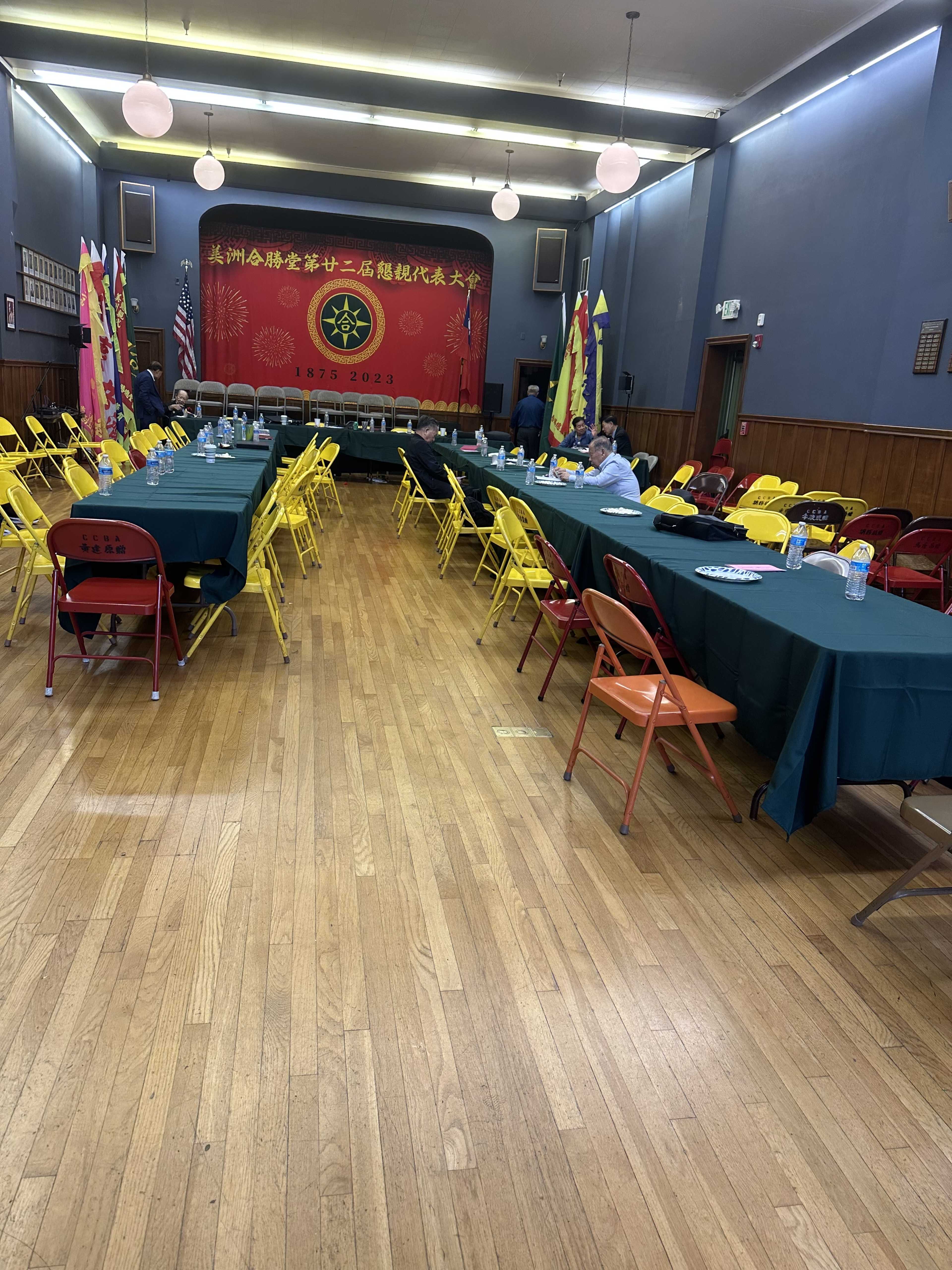 A spacious hall features rows of tables with yellow chairs, a red backdrop decorated with flags, and an empty stage at the front.