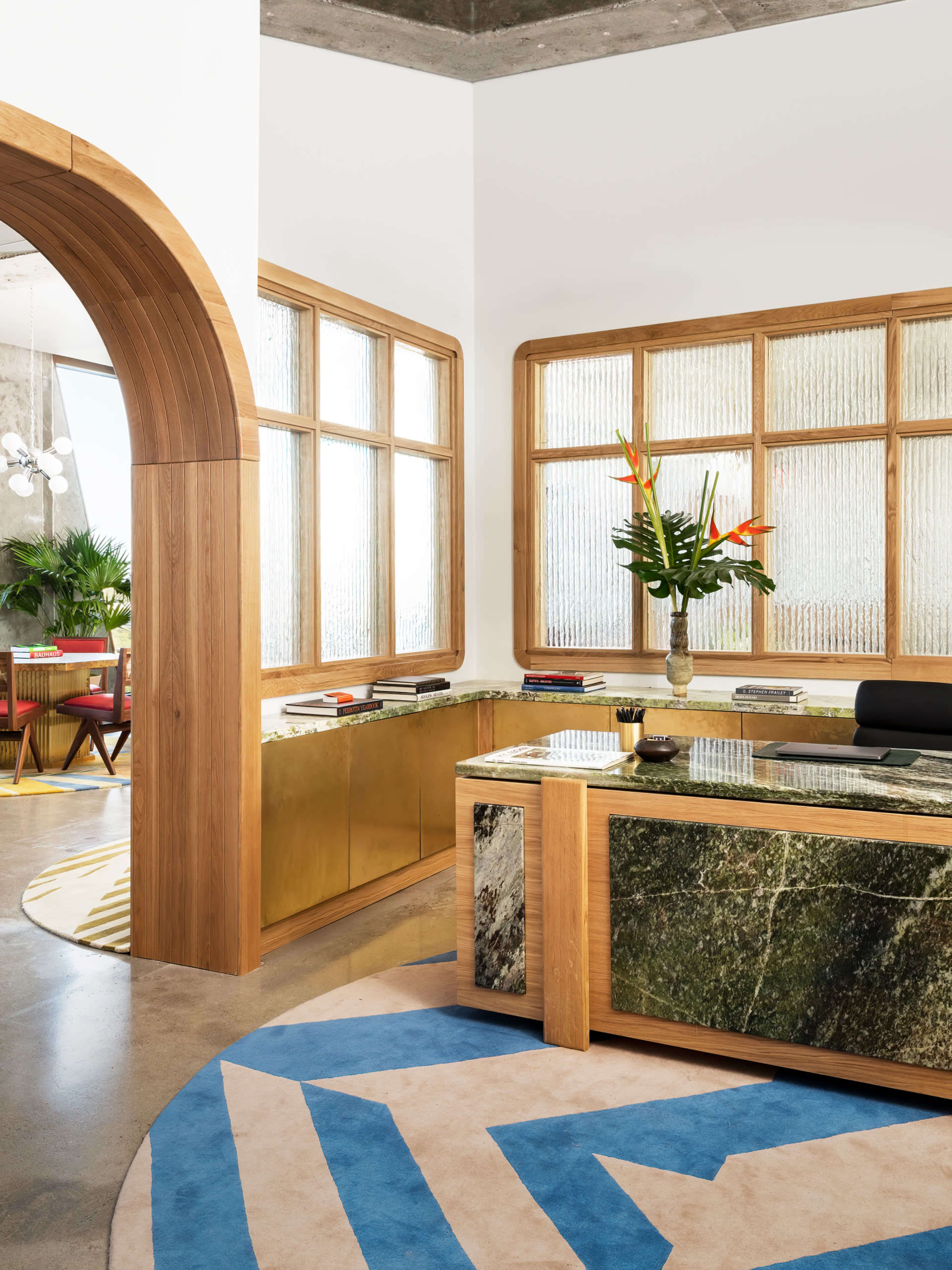 A modern office interior featuring a sleek reception desk made of green marble and brass, large windows with textured glass, and a vibrant area rug with geometric patterns.