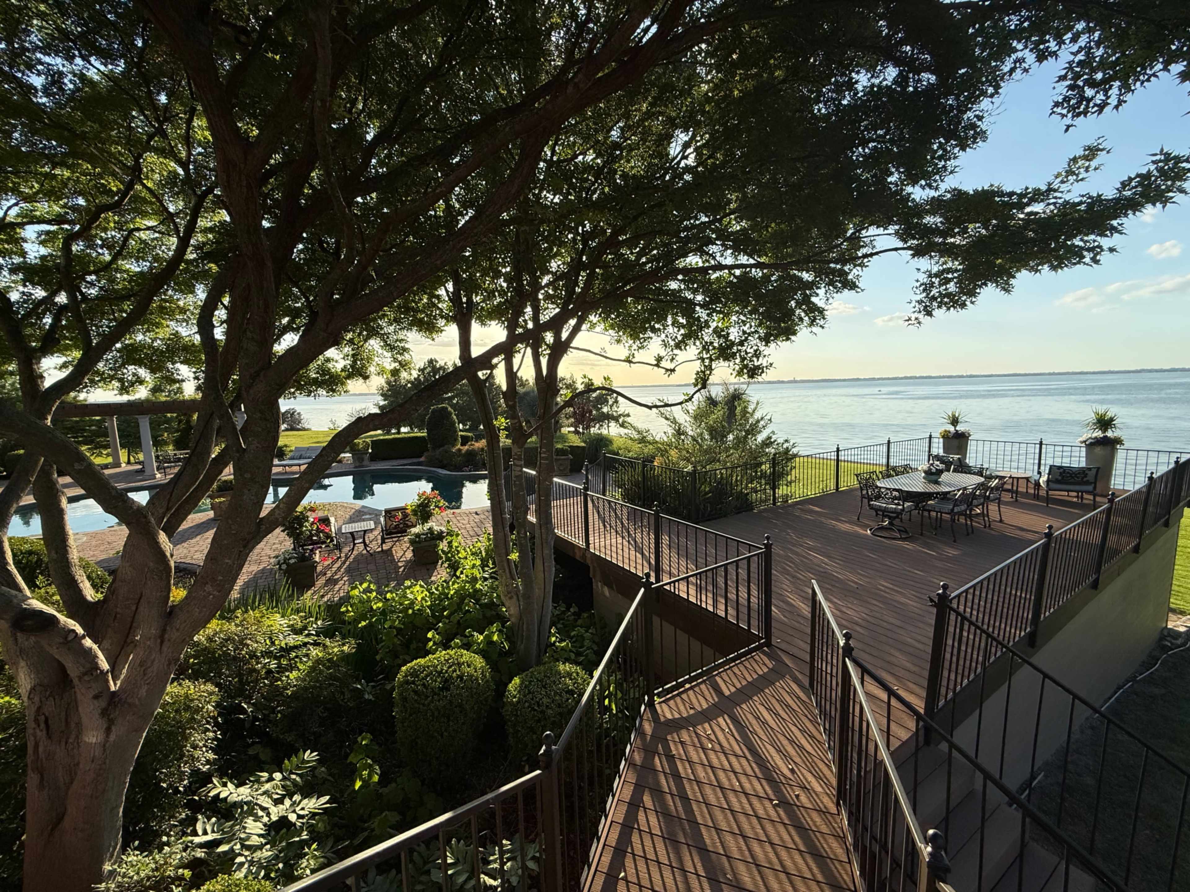 The image shows a well-maintained outdoor deck overlooking a pool and lake, surrounded by lush greenery and landscaped gardens.