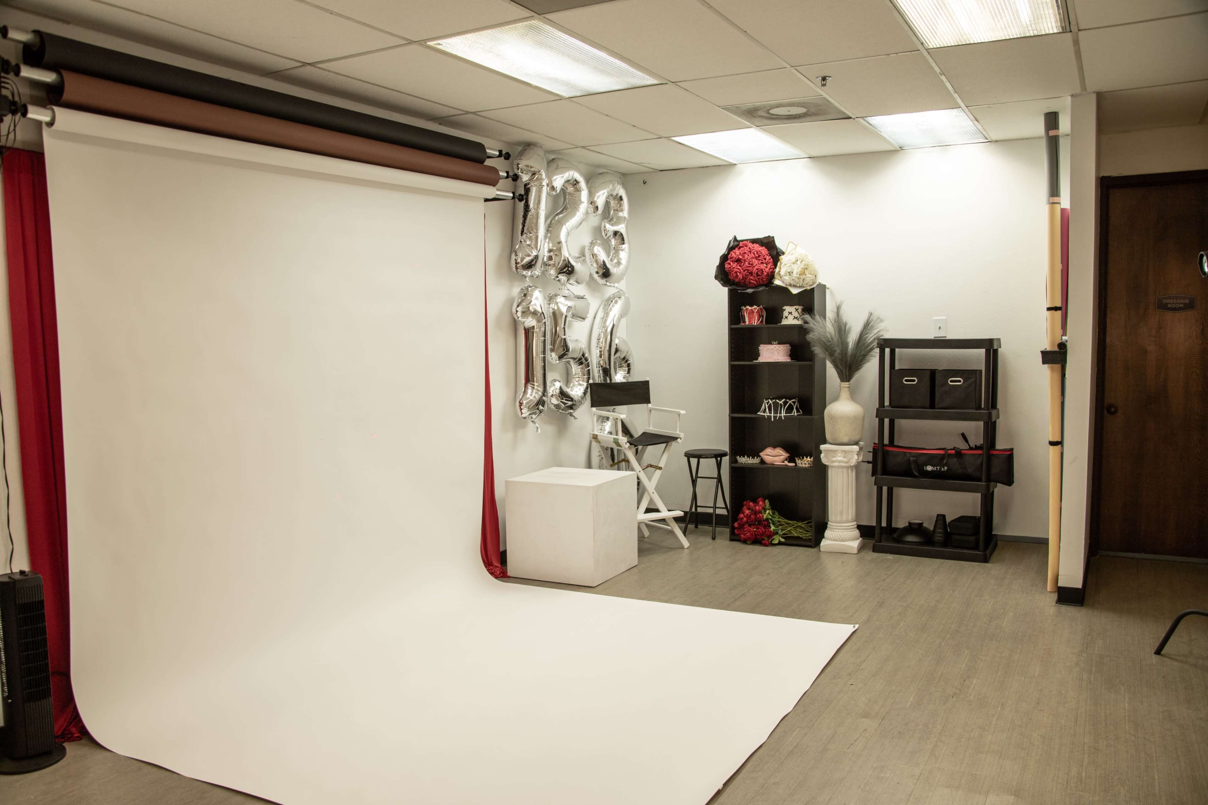 The image shows a photography studio equipped with a plain white backdrop, a styling area with decorative elements, and a shelving unit filled with props.
