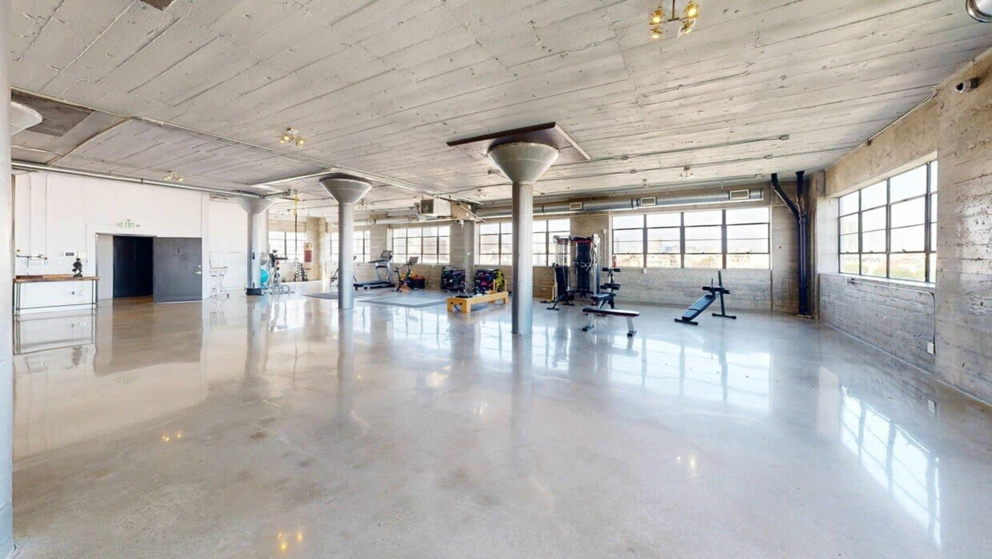 The image shows a spacious gym area with large windows, polished concrete floors, and various exercise equipment arranged throughout.