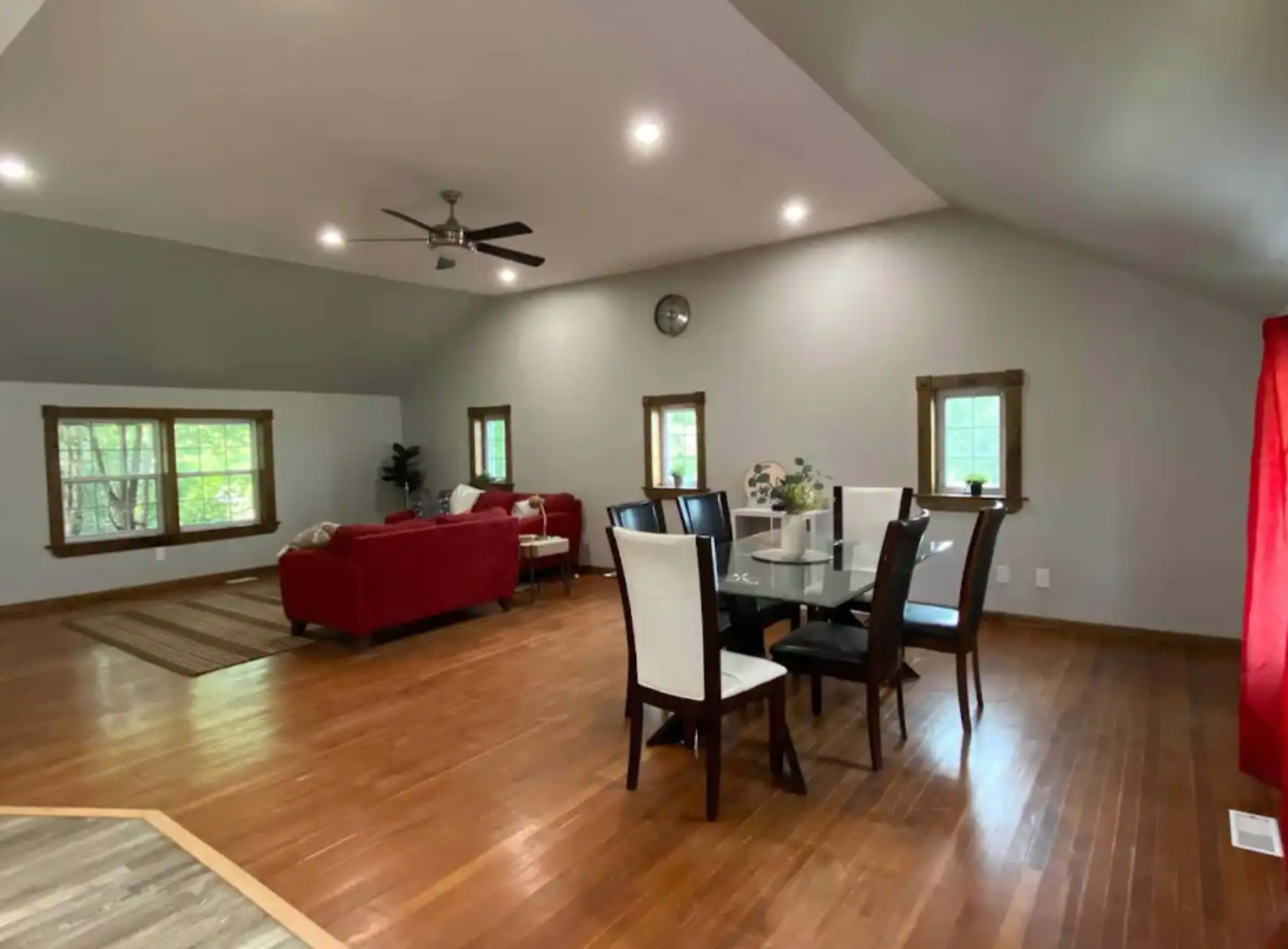 The image shows a spacious room with hardwood flooring, featuring a dining area with a table and chairs and a living area with a red sofa and windows.