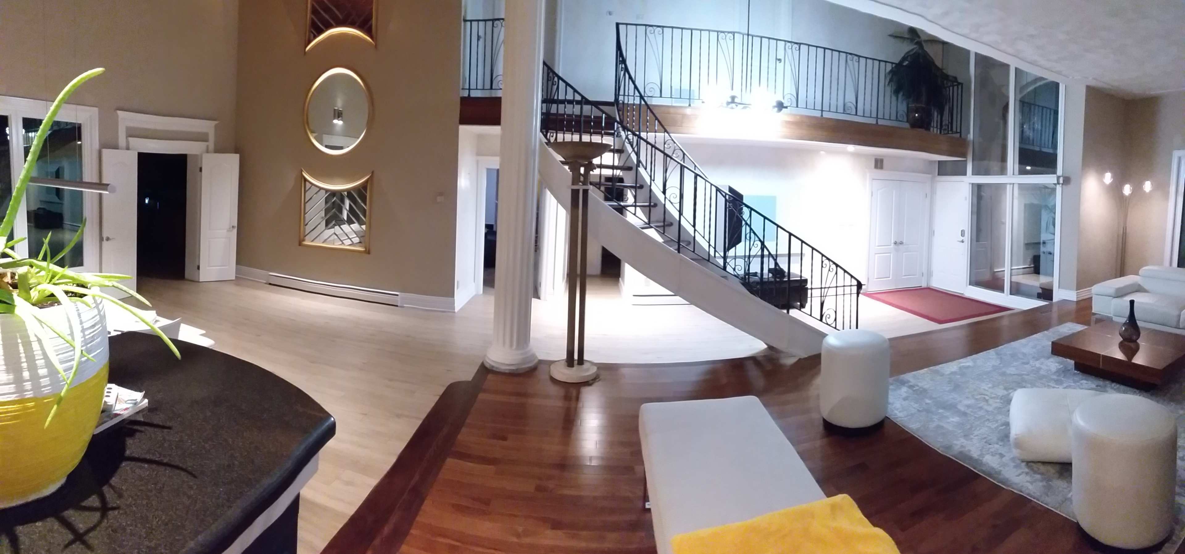 A spacious living area features a curved staircase, large windows, and modern furnishings arranged around a central rug.