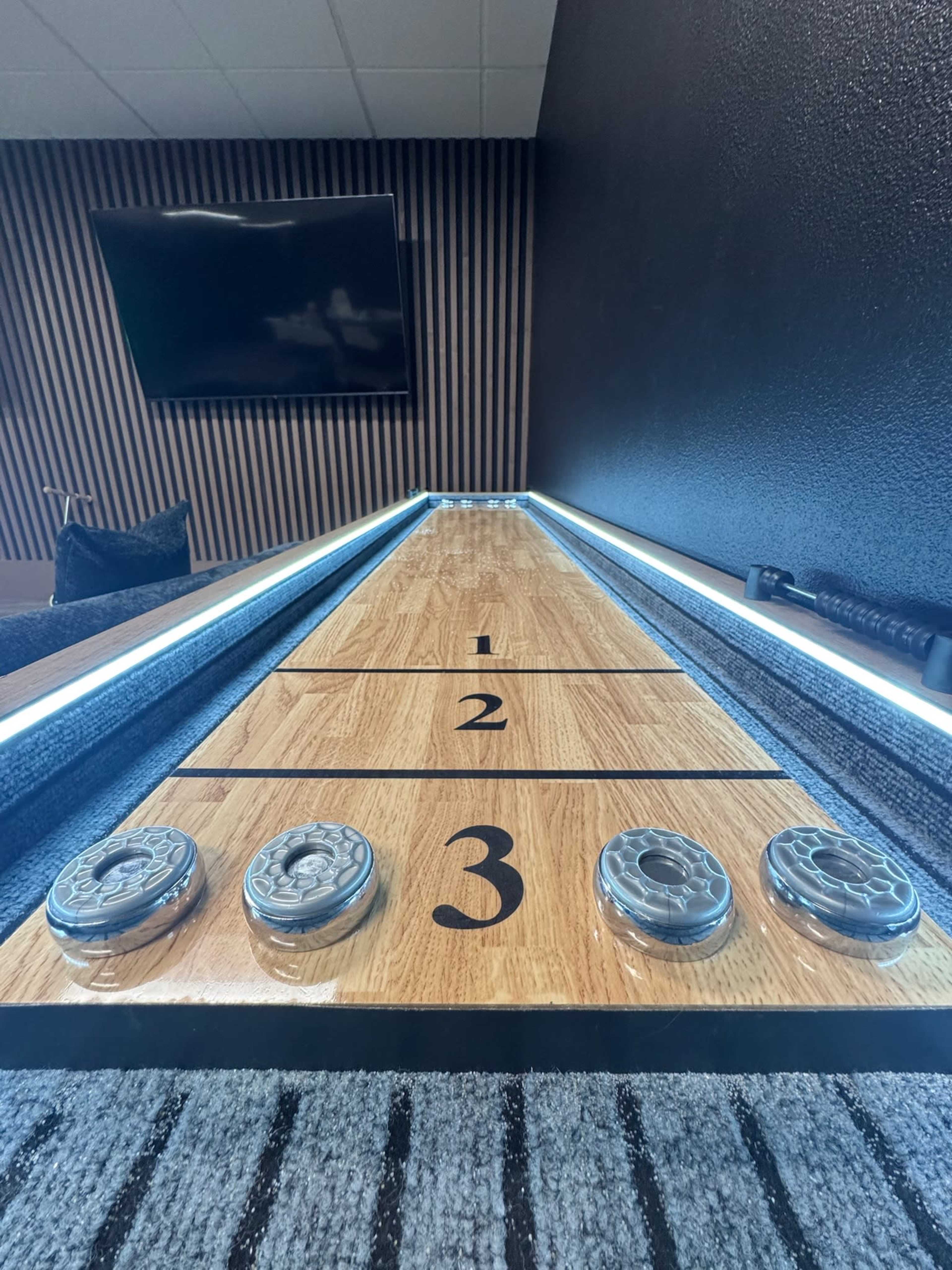 The image shows a wooden shuffleboard court with scoring sections labeled one, two, and three, along with pucks positioned at the end of the court.