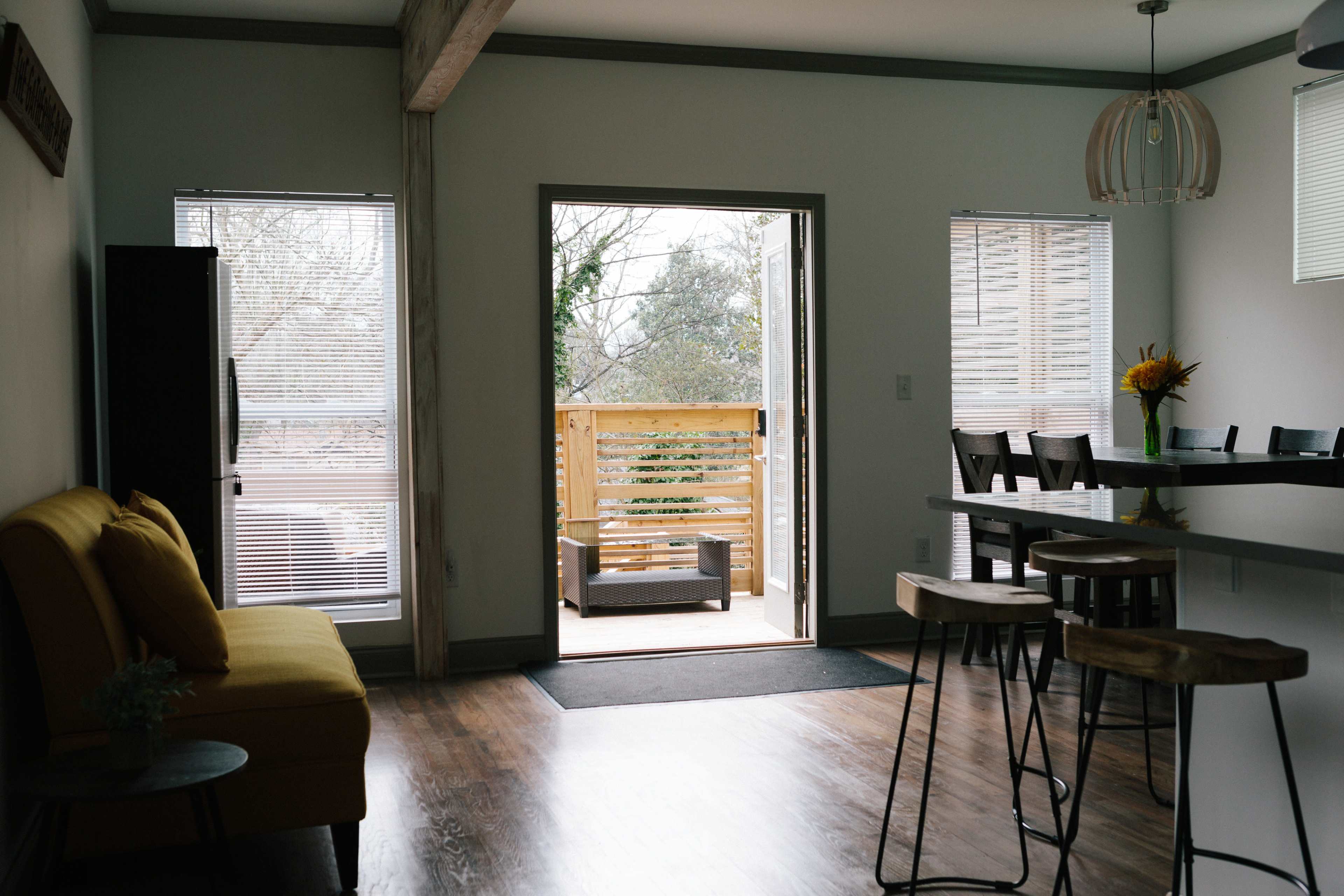 A bright, open living space with a yellow sofa, a kitchen area with bar stools, and large windows looking out onto a balcony.