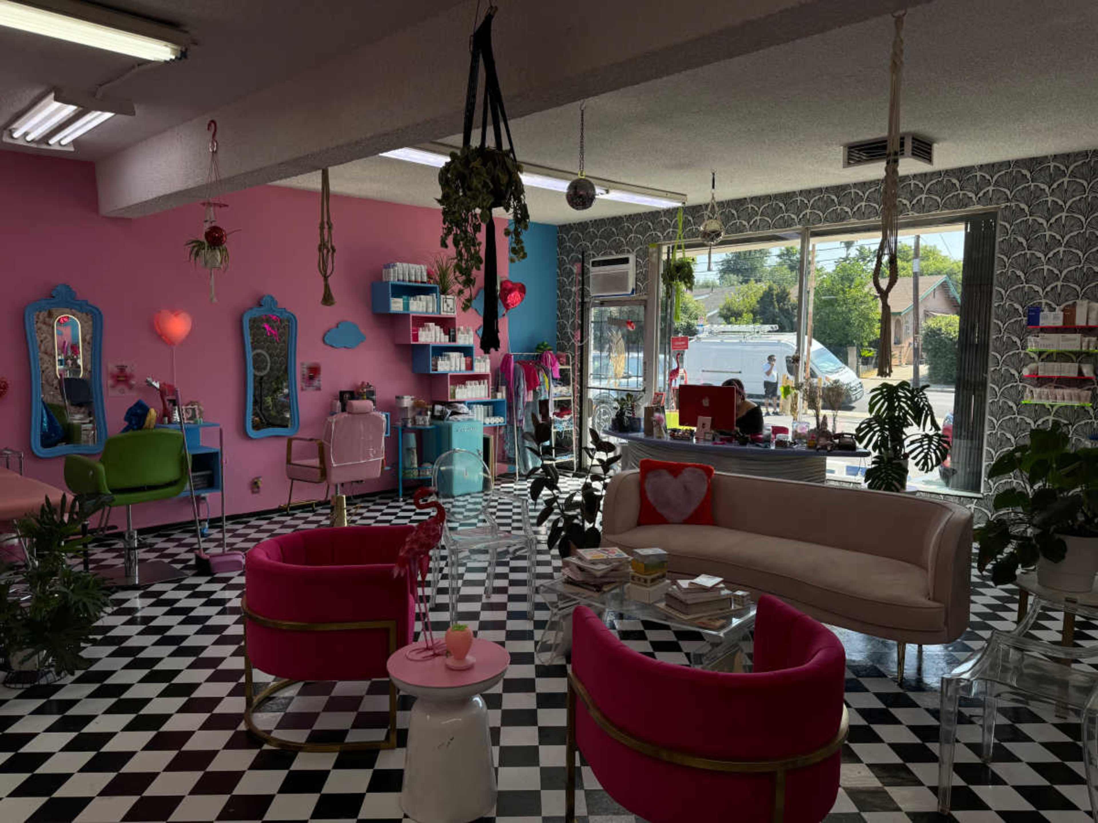 The image shows a brightly decorated salon with pink walls, black and white checkered flooring, and a variety of seating and decorative plants.