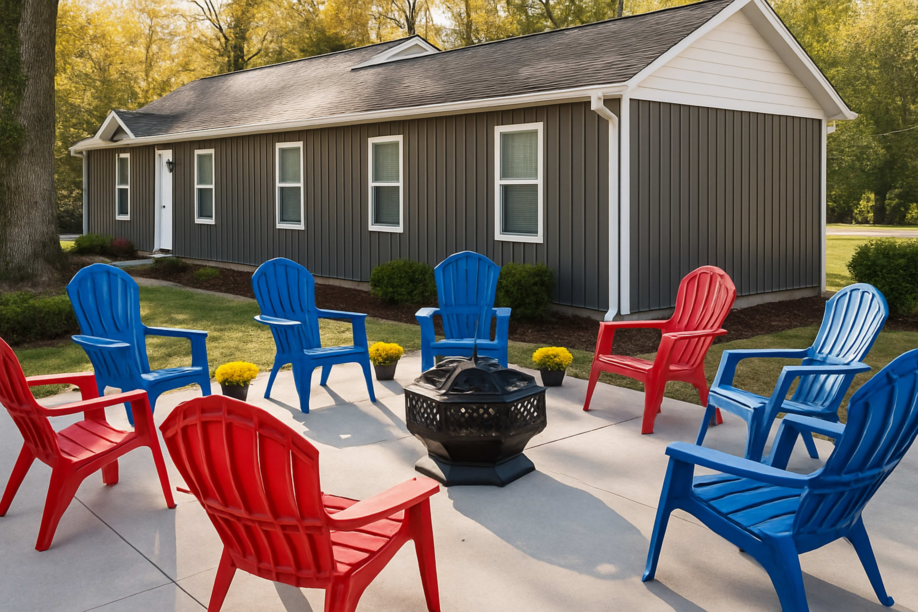 A circular arrangement of red and blue Adirondack chairs surrounds a fire pit in front of a gray house with a white roof.