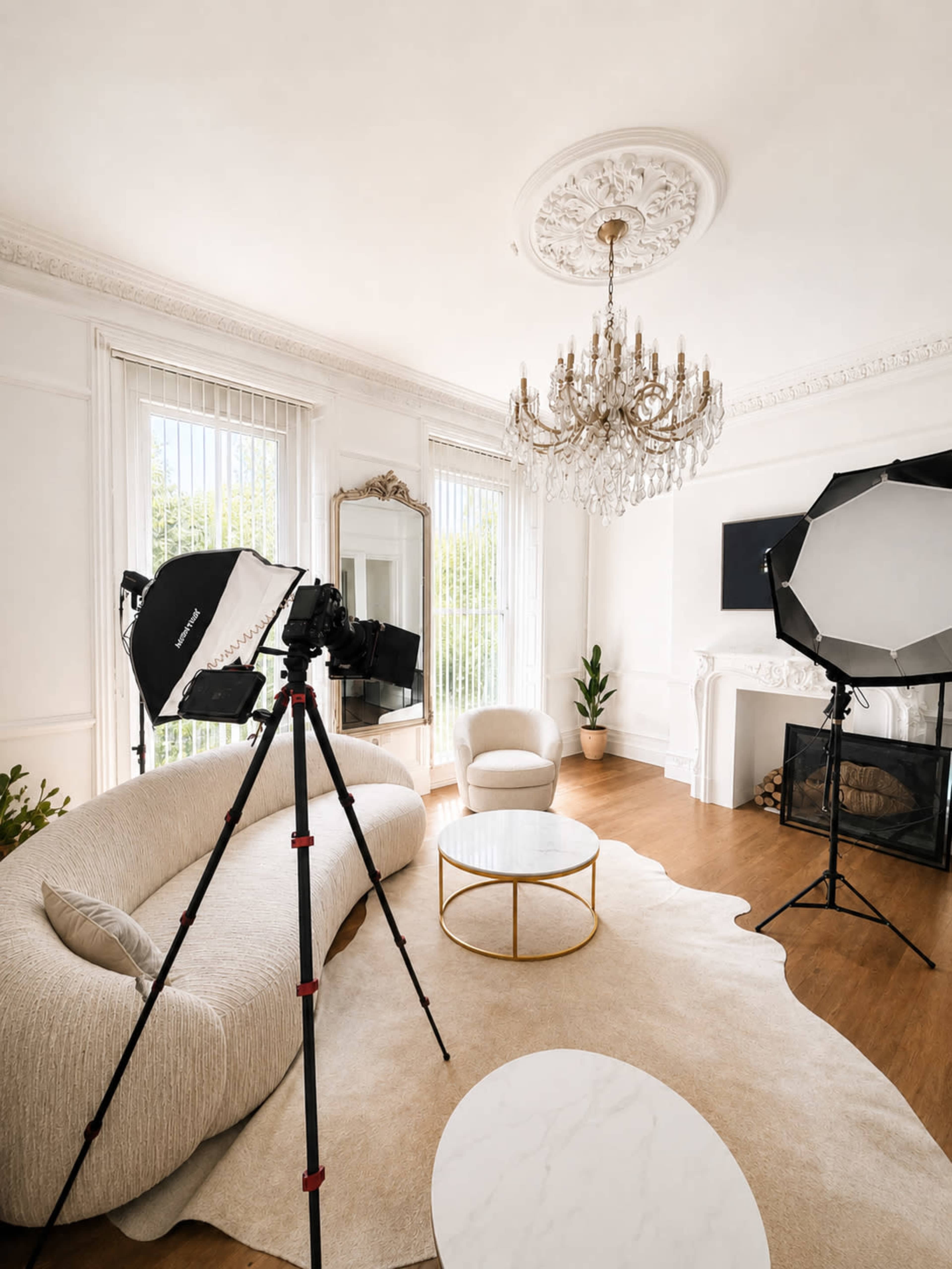 A well-lit photography studio features a soft sofa, a round coffee table, a chandelier, and professional lighting equipment set up against a backdrop of large windows.