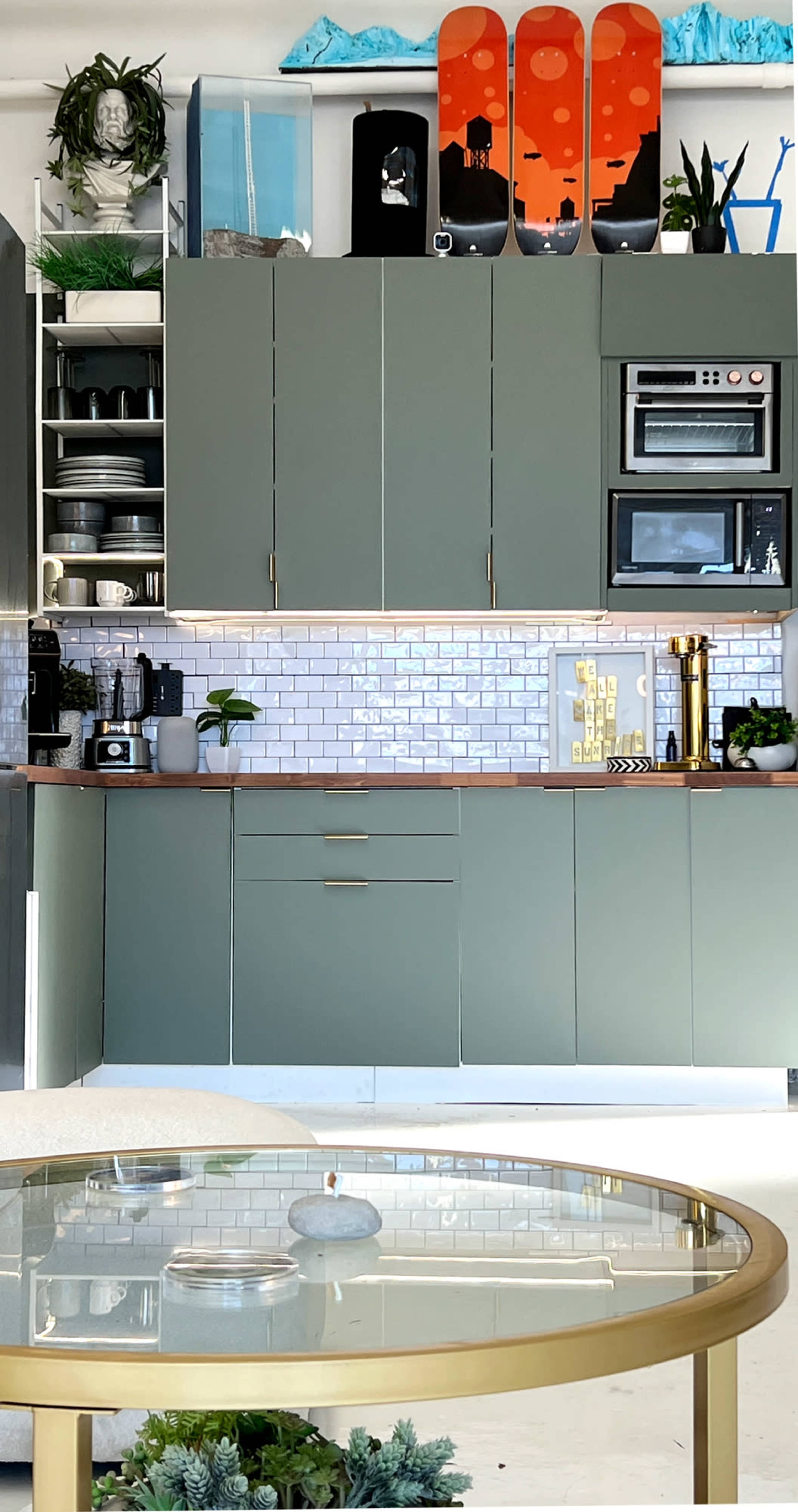 The image shows a modern kitchen featuring green cabinets, open shelves with kitchen items, and a round glass dining table with a gold base.
