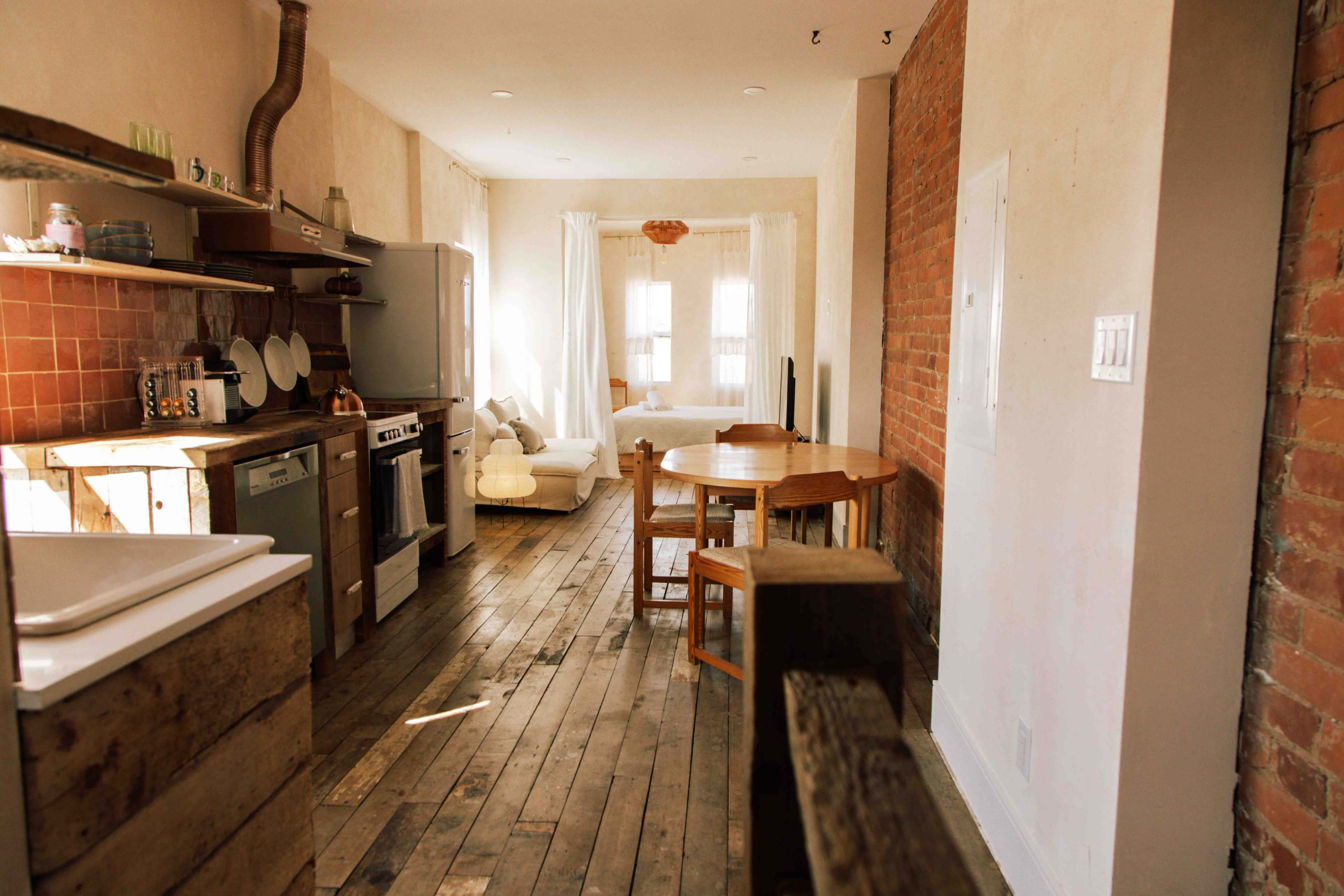 The image shows a rustic kitchen area with wooden floors, exposed brick walls, and a dining table set up in a brightly lit space.