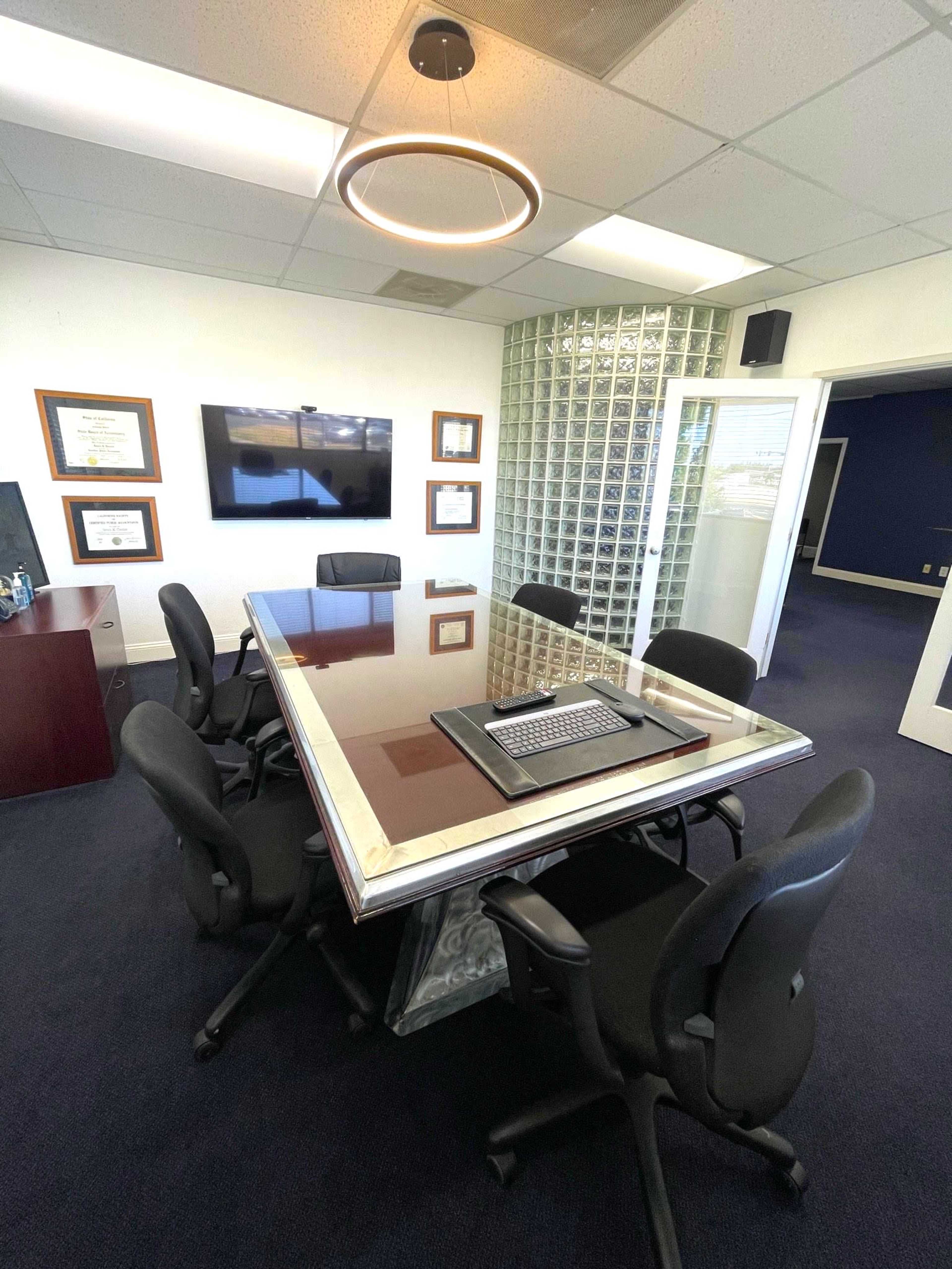 A conference room featuring a large table surrounded by black chairs, with a wall of glass bricks and a television mounted on the wall.