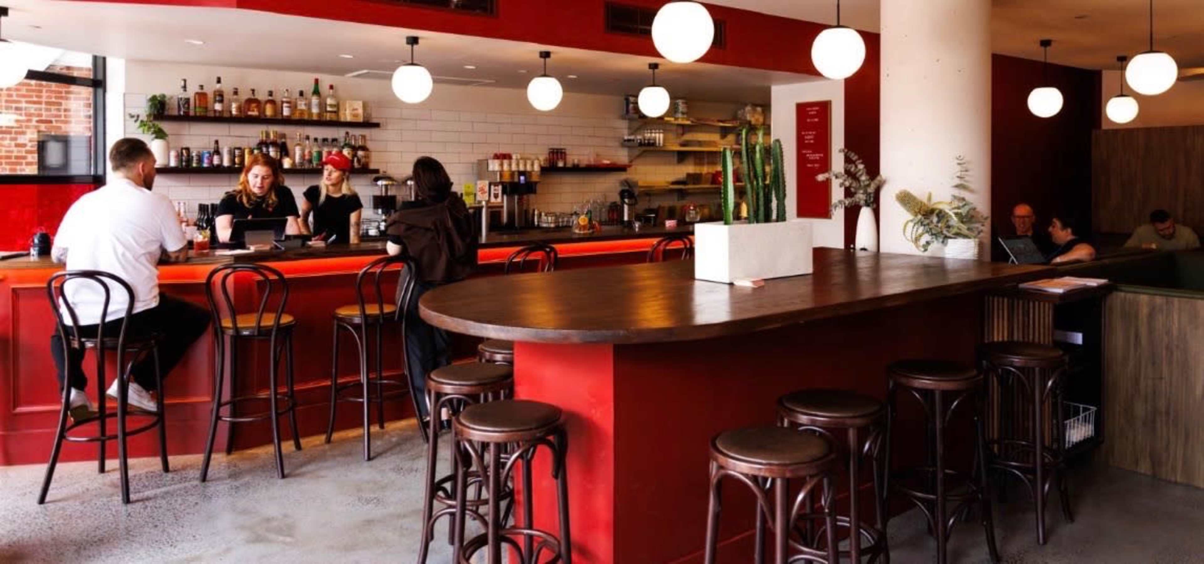 The image shows a modern cafe interior featuring a long wooden bar, string pendant lights, and a mix of patrons seated at the bar and tables.