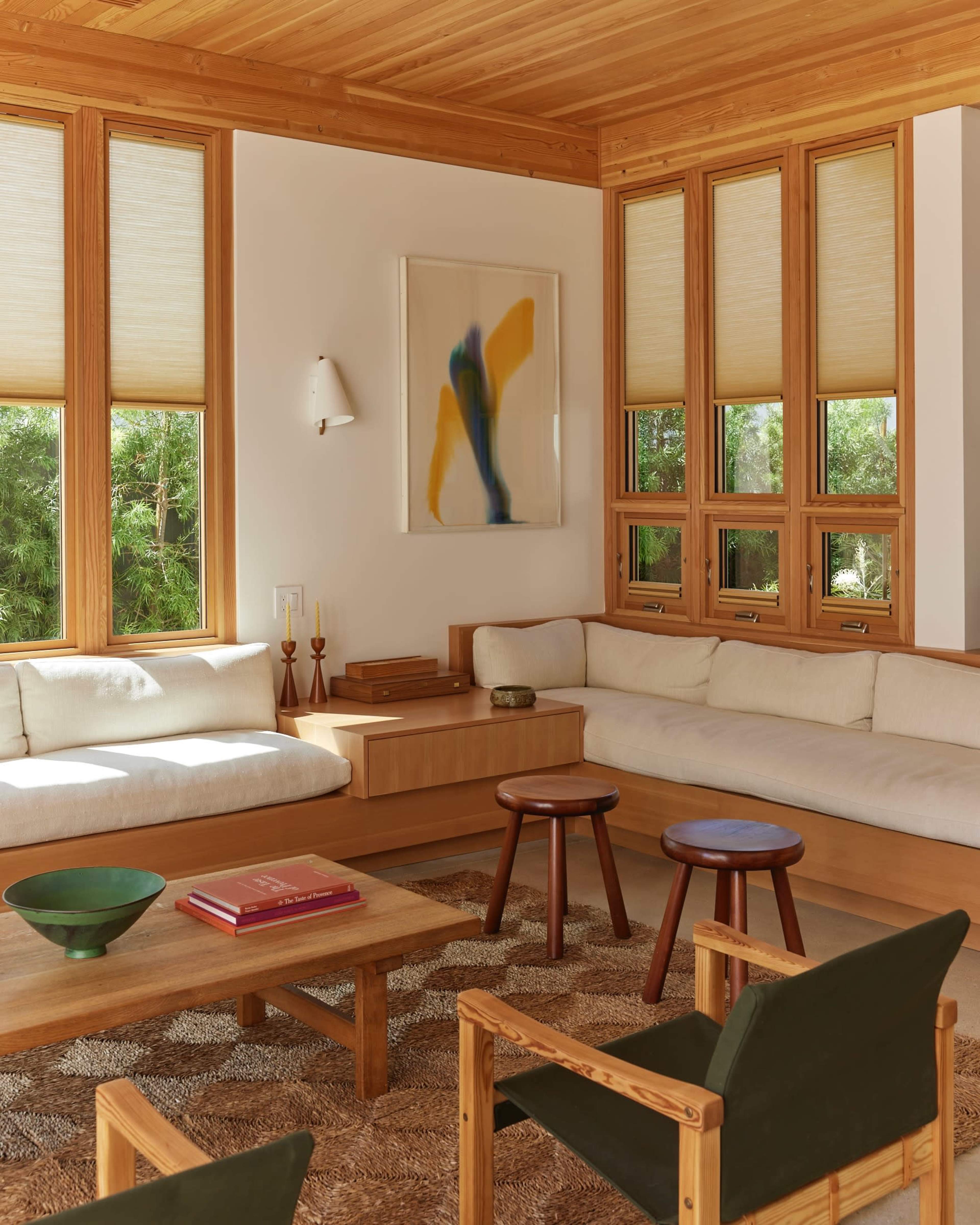 A modern living room with wooden walls and furniture, featuring a sectional couch, a coffee table, and large windows with light-filtering shades.