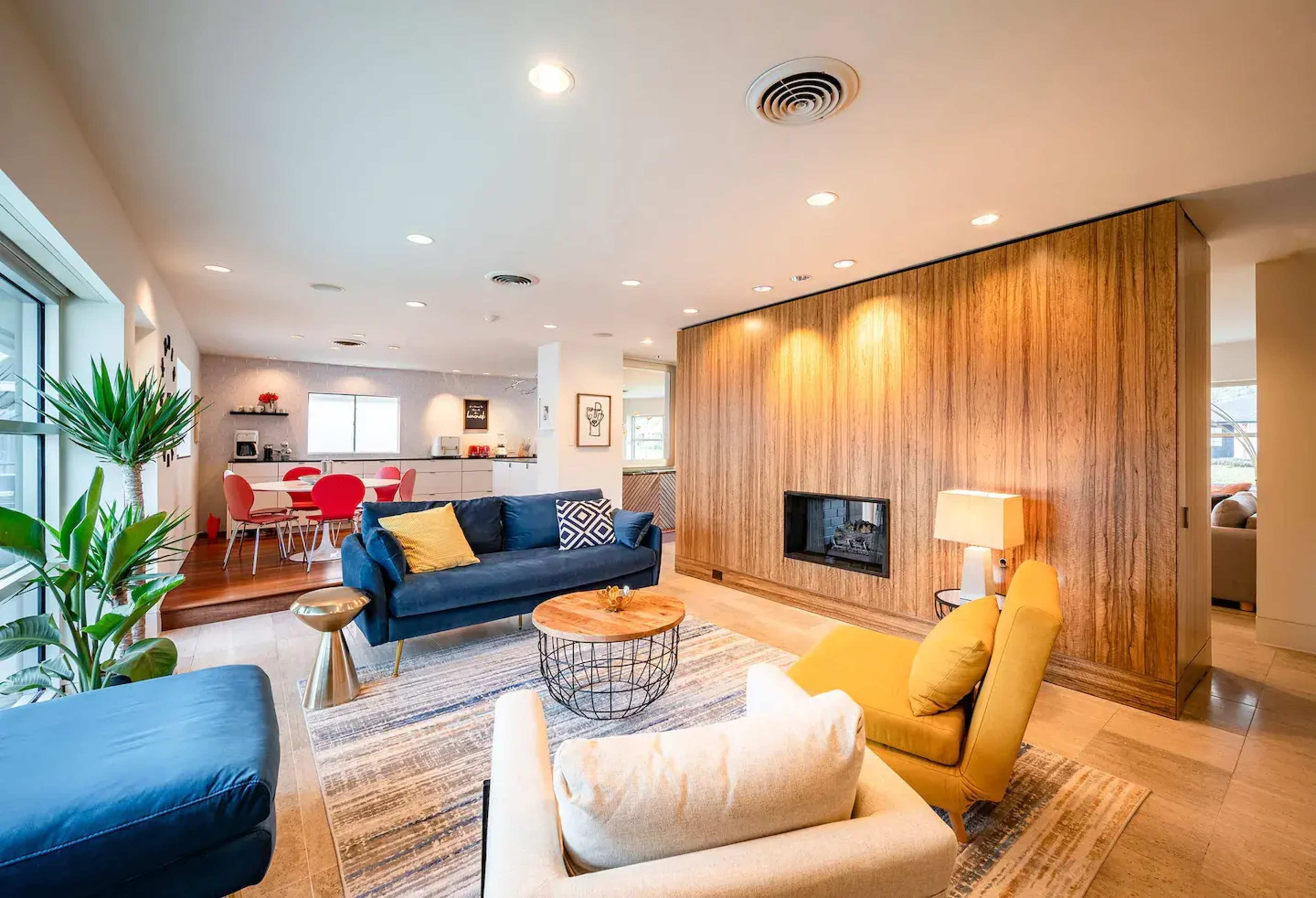 A modern living room features two sofas, a coffee table, and a wall-mounted fireplace, with a dining area visible in the background.