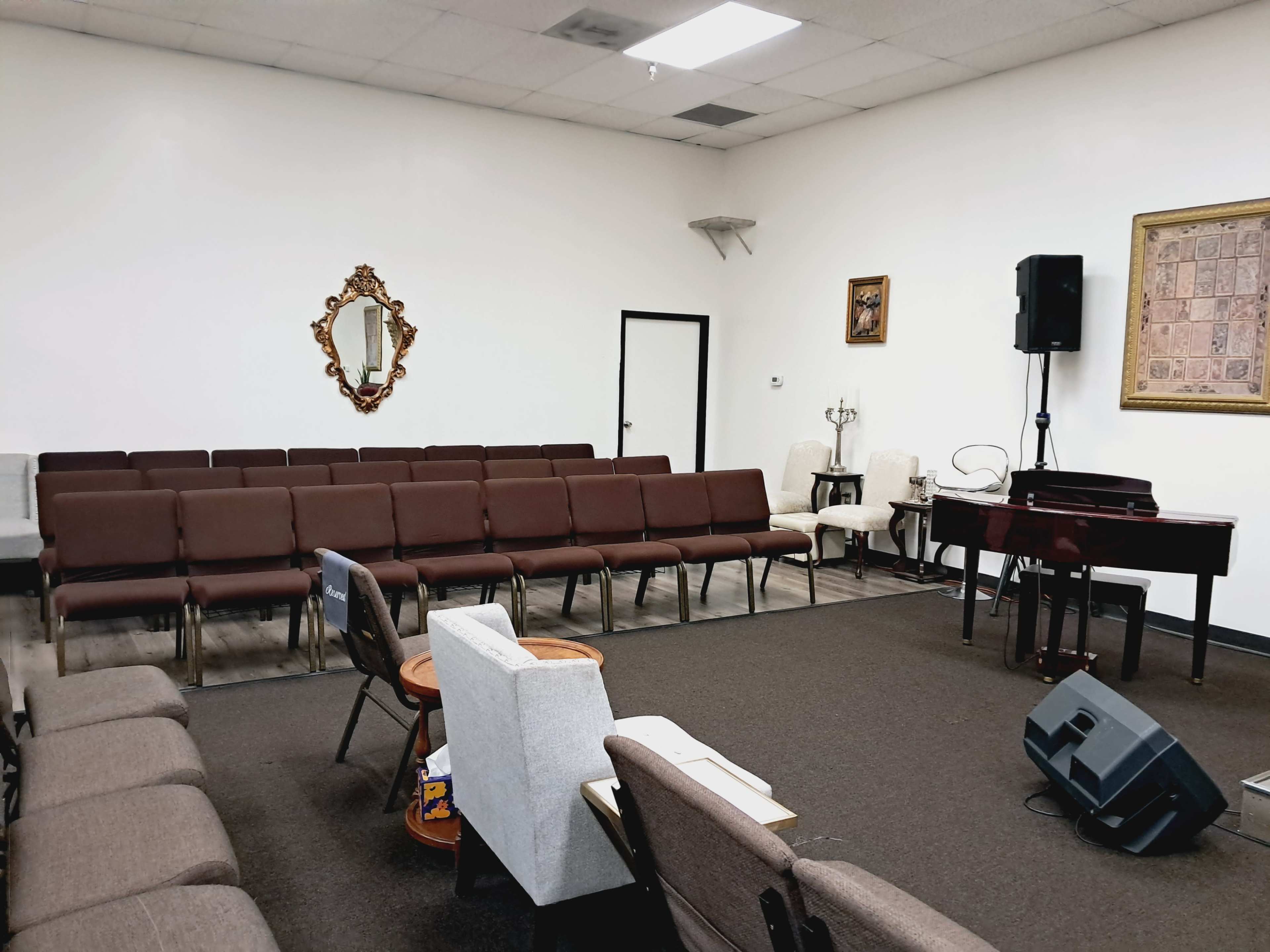 The image shows a small indoor space with rows of brown chairs facing a piano and a speaker system, accented by a mirror and framed artwork on the walls.