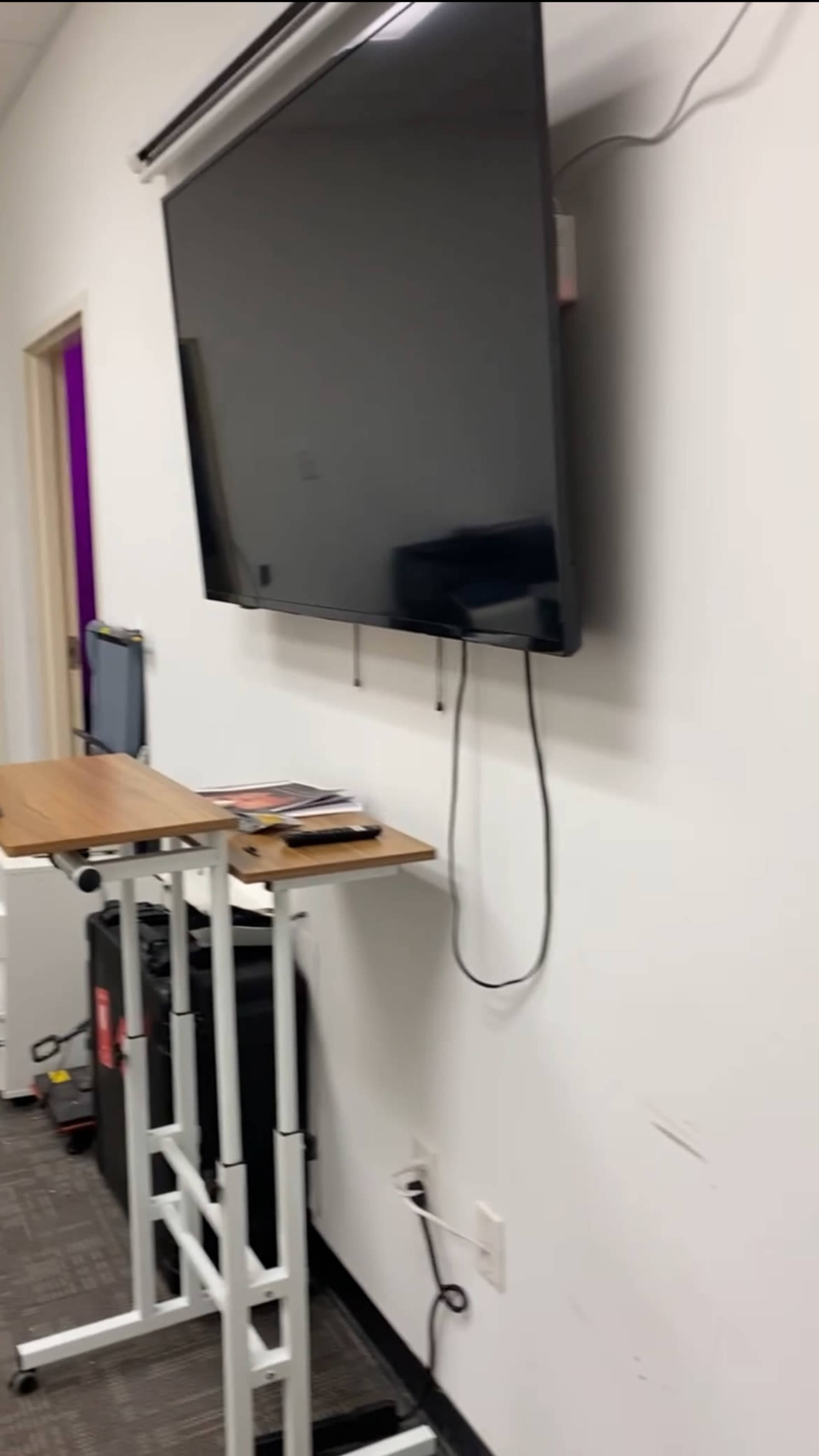 A wall-mounted television above a small wooden table in a room with a gray carpet and white walls.