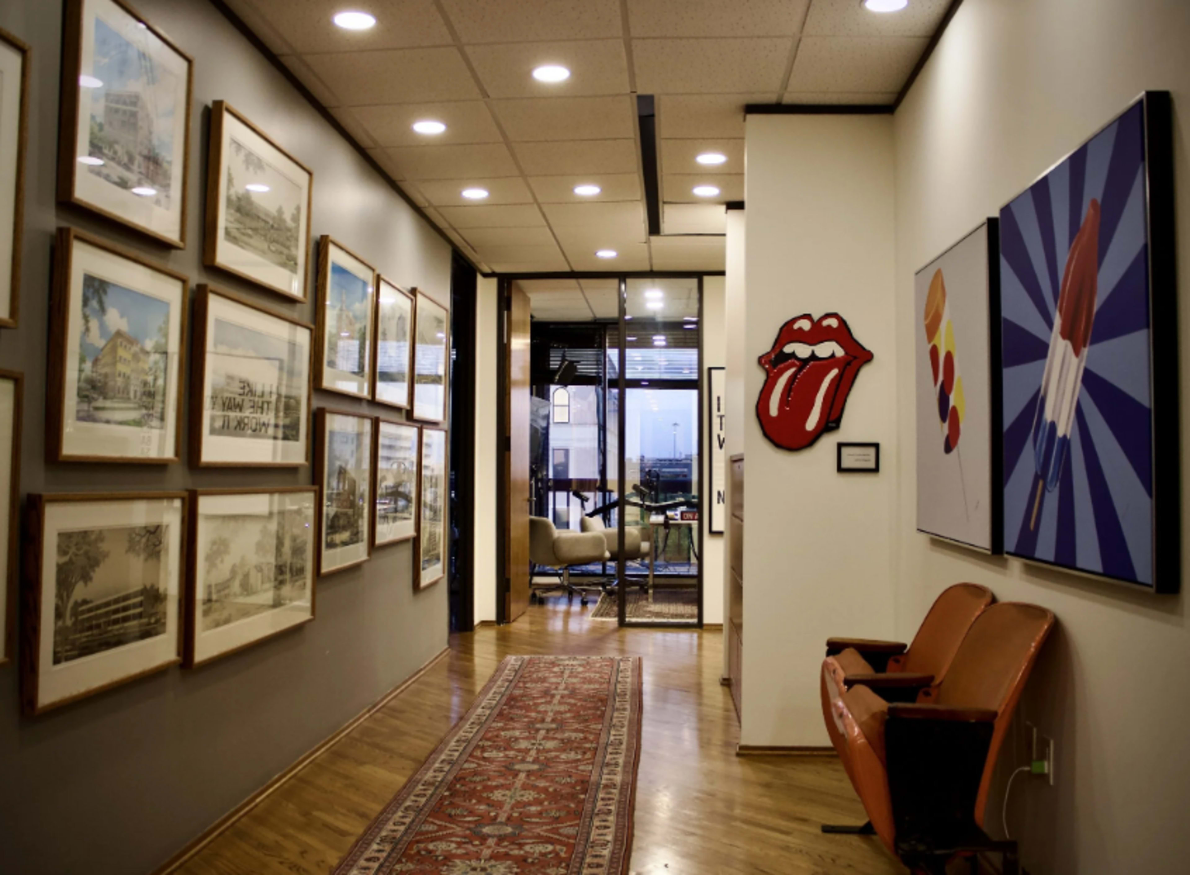 A hallway features framed artwork on the walls, with a red sofa at one end and an office space visible in the background.