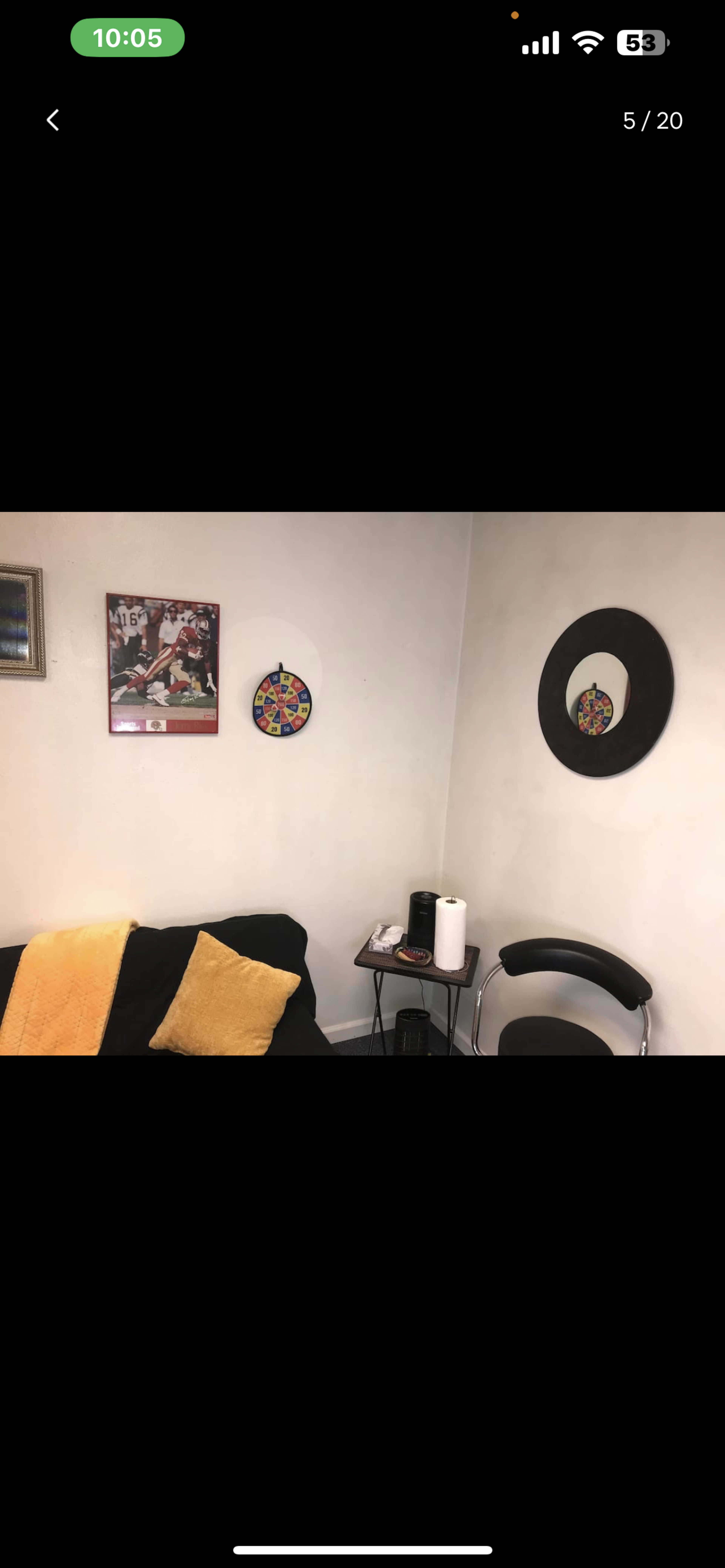 The image displays a corner of a room featuring a black couch with a yellow throw, two wall decorations, a small round table, and a chair.