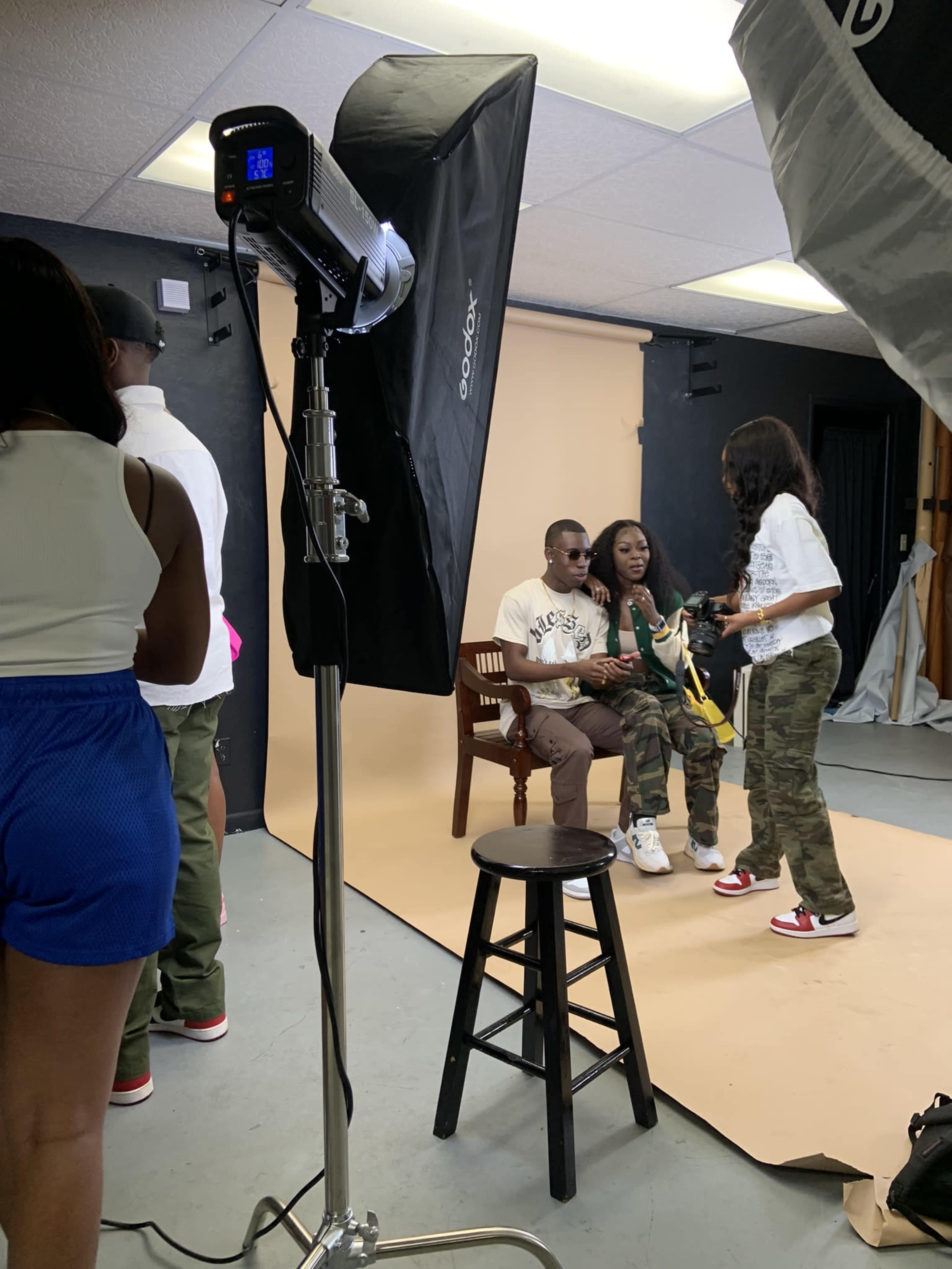 A photoshoot takes place in a studio with subjects posing on a chair against a backdrop, while a photographer and assistants manage lighting and setup.