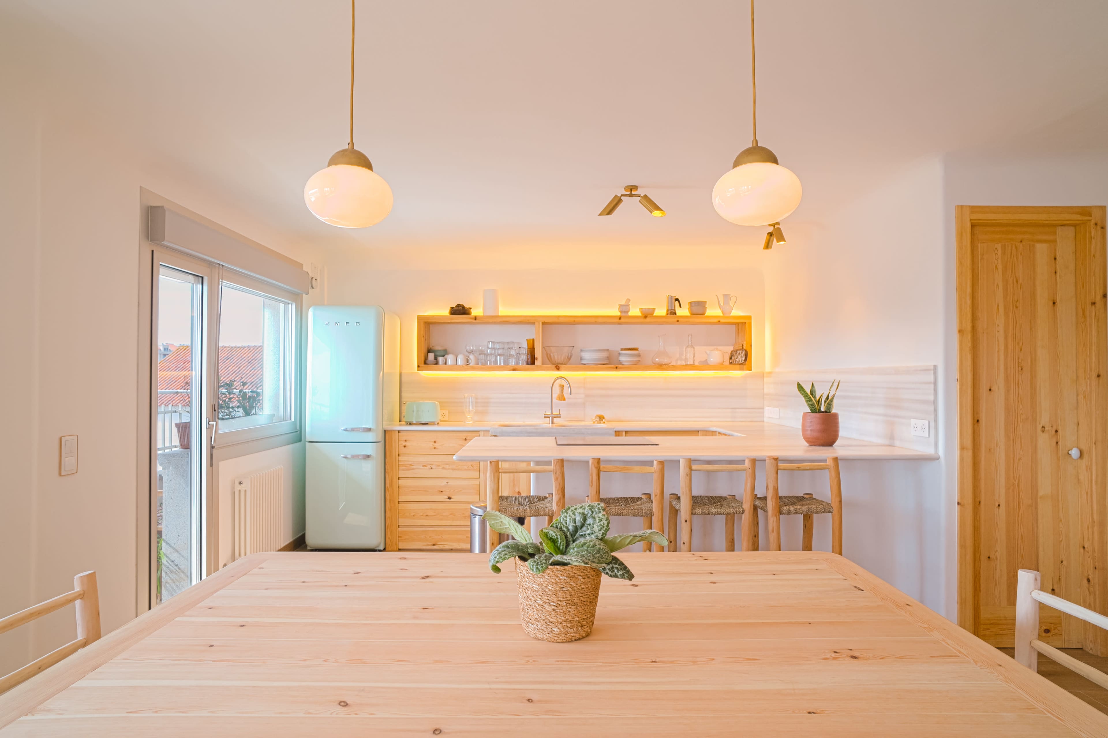 A modern kitchen features a wooden dining table, a bright green refrigerator, and pendant lights illuminating a minimalist countertop with plants and kitchenware.