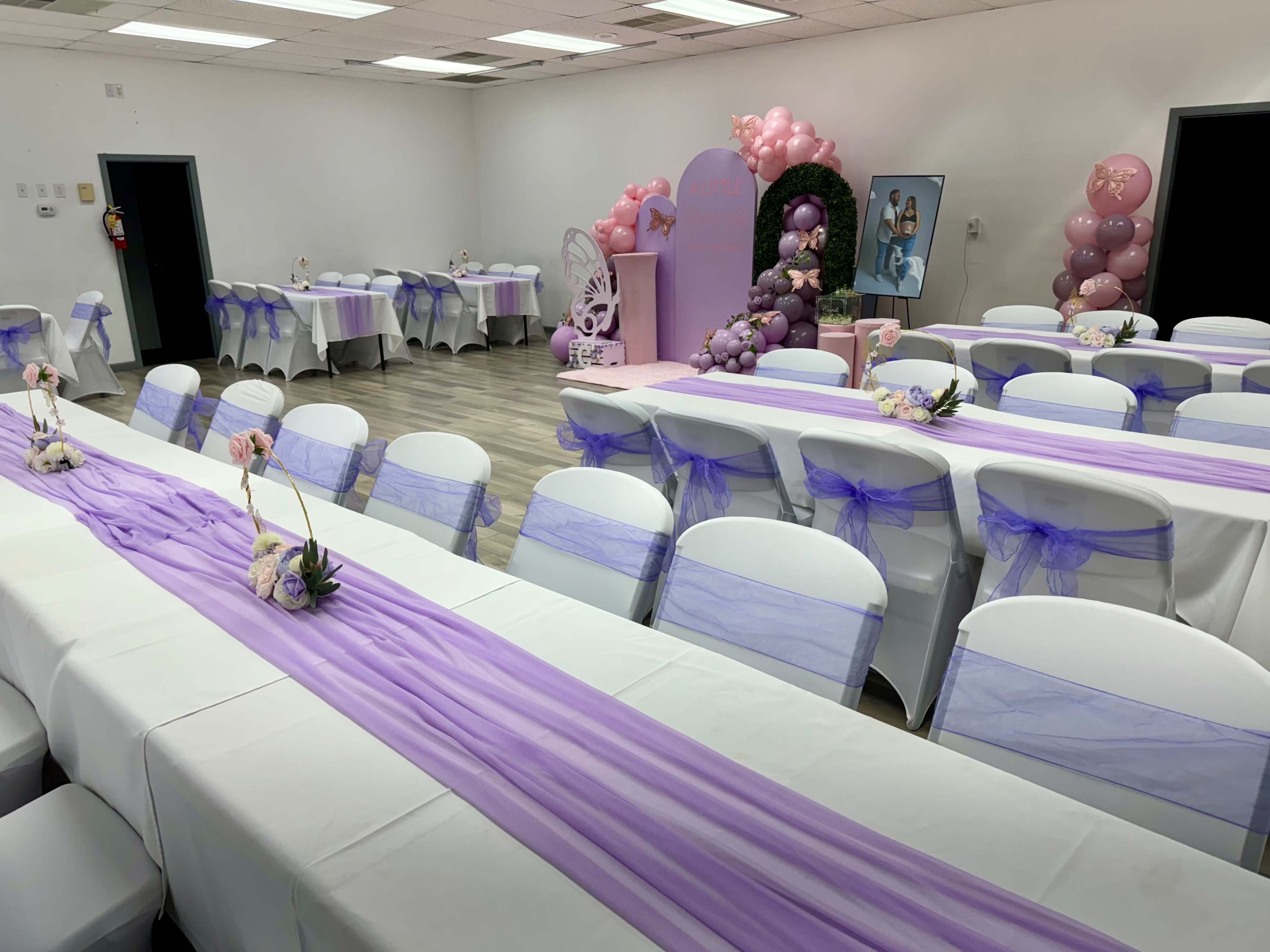 A banquet hall decorated with purple table covers and floral centerpieces, featuring balloons and a photo display at one end.