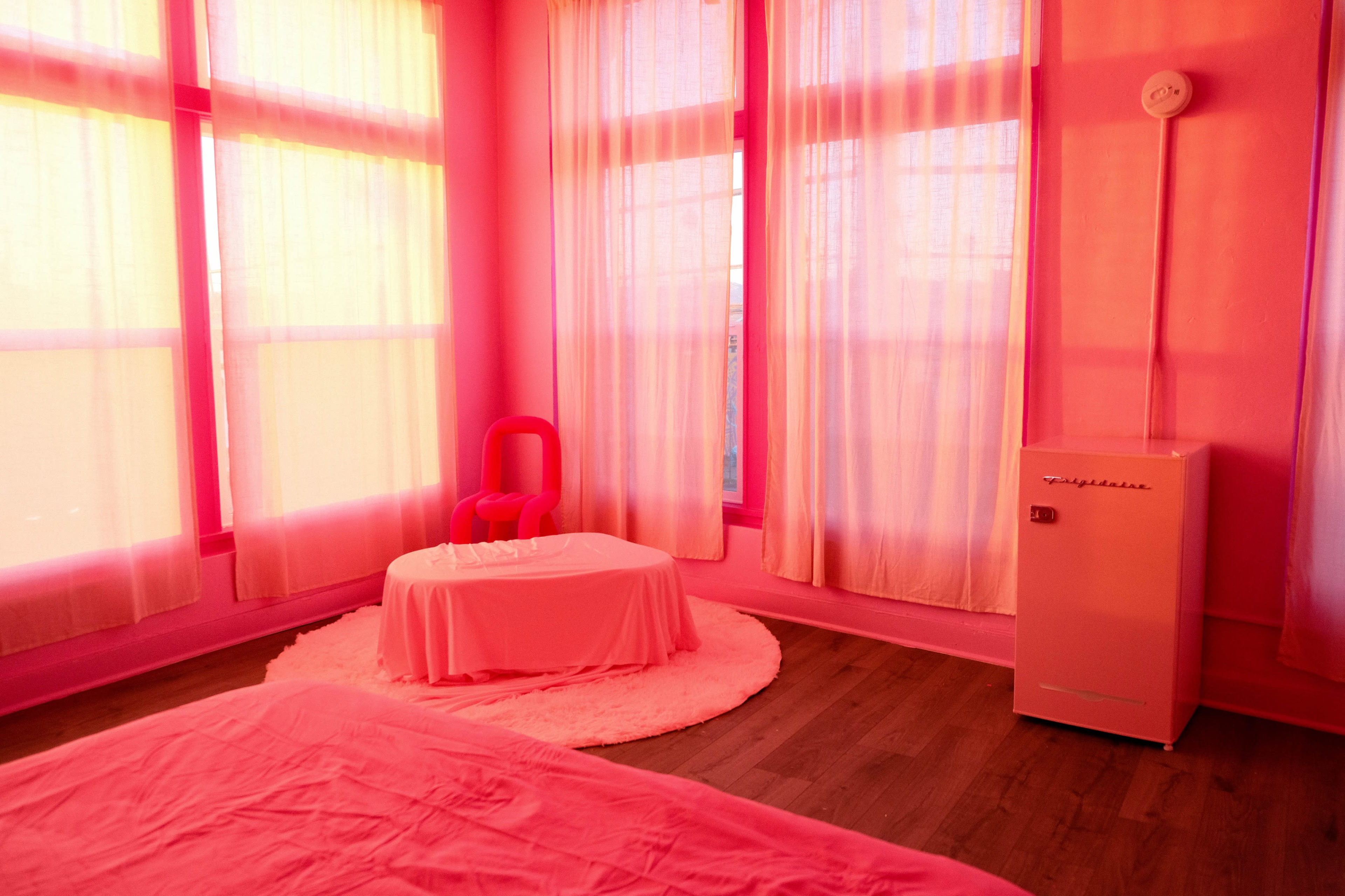 A room with pink walls, large windows covered with sheer pink curtains, a white tablecloth draped over a small table, and a modern white refrigerator.