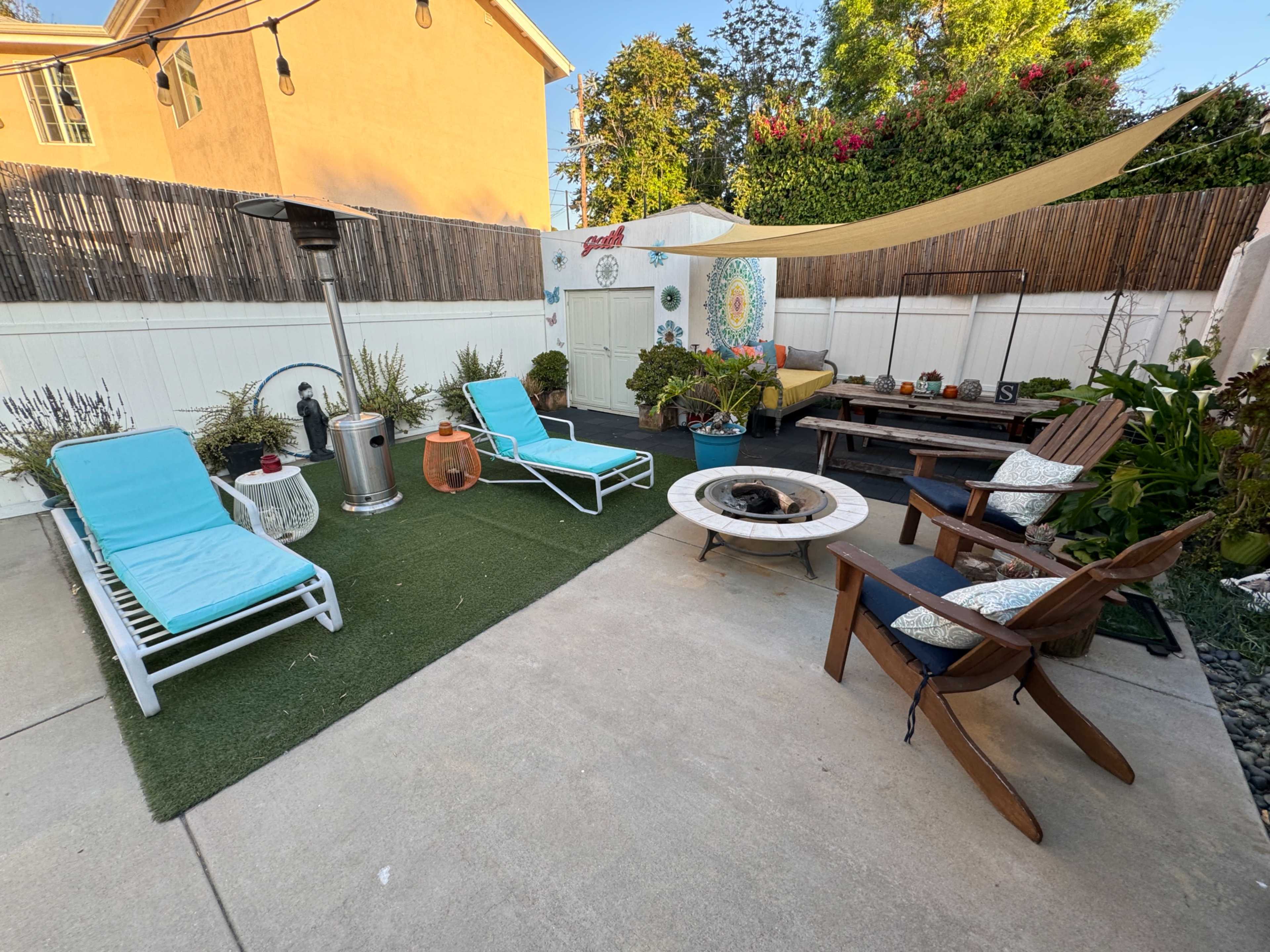 The scene features a landscaped backyard with lounge chairs, a fire pit, and various planters, set against a backdrop of a small storage shed and wooden fencing.