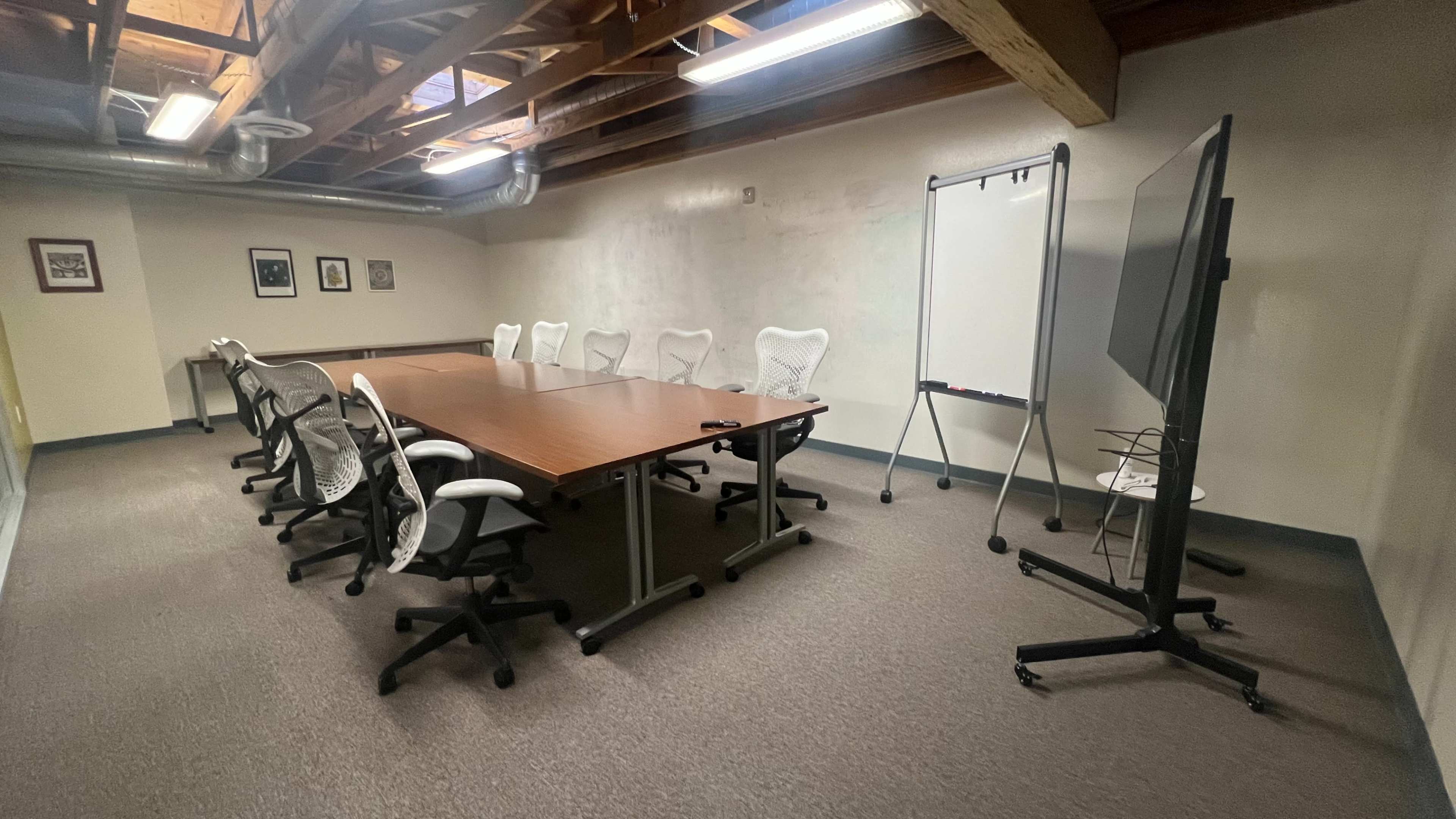 A conference room features a long wooden table surrounded by eight chairs, a whiteboard on a stand, and a large display screen.