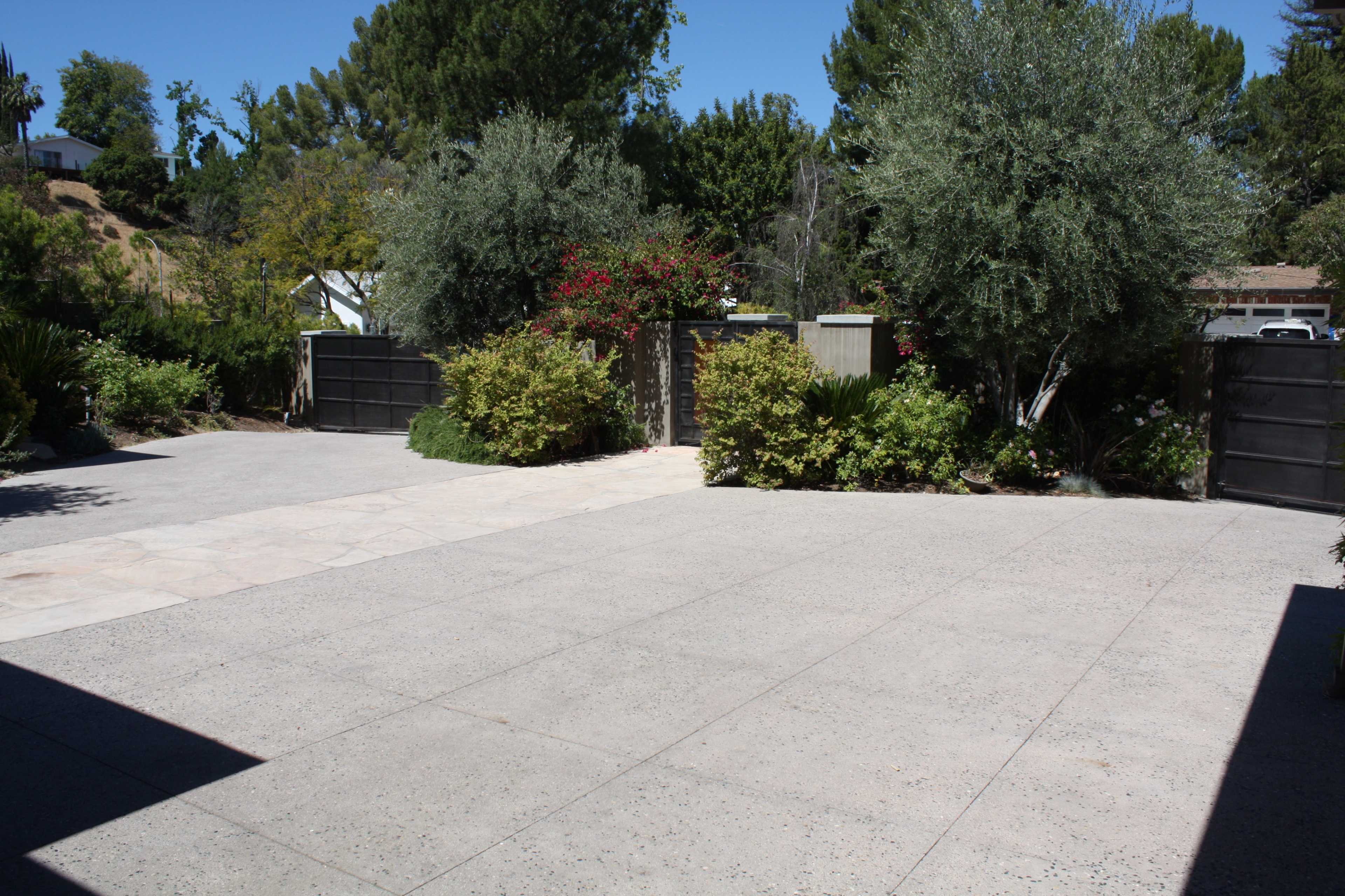 A landscaped outdoor area with a paved driveway, surrounded by greenery and a pair of gates in the background.