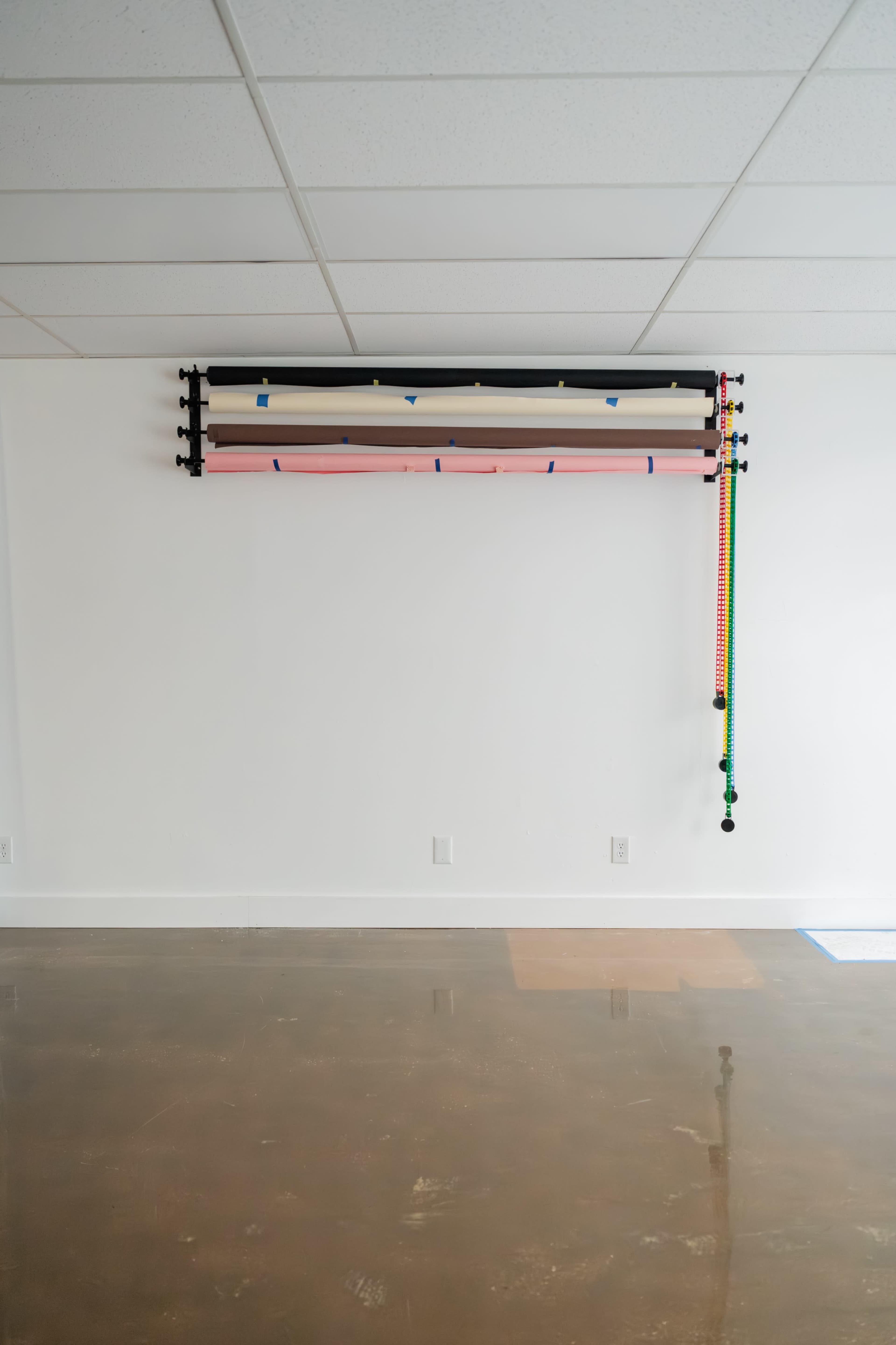 The image shows a wall with multiple rolls of backdrop paper and colorful straps mounted above, alongside a smooth, reflective floor.