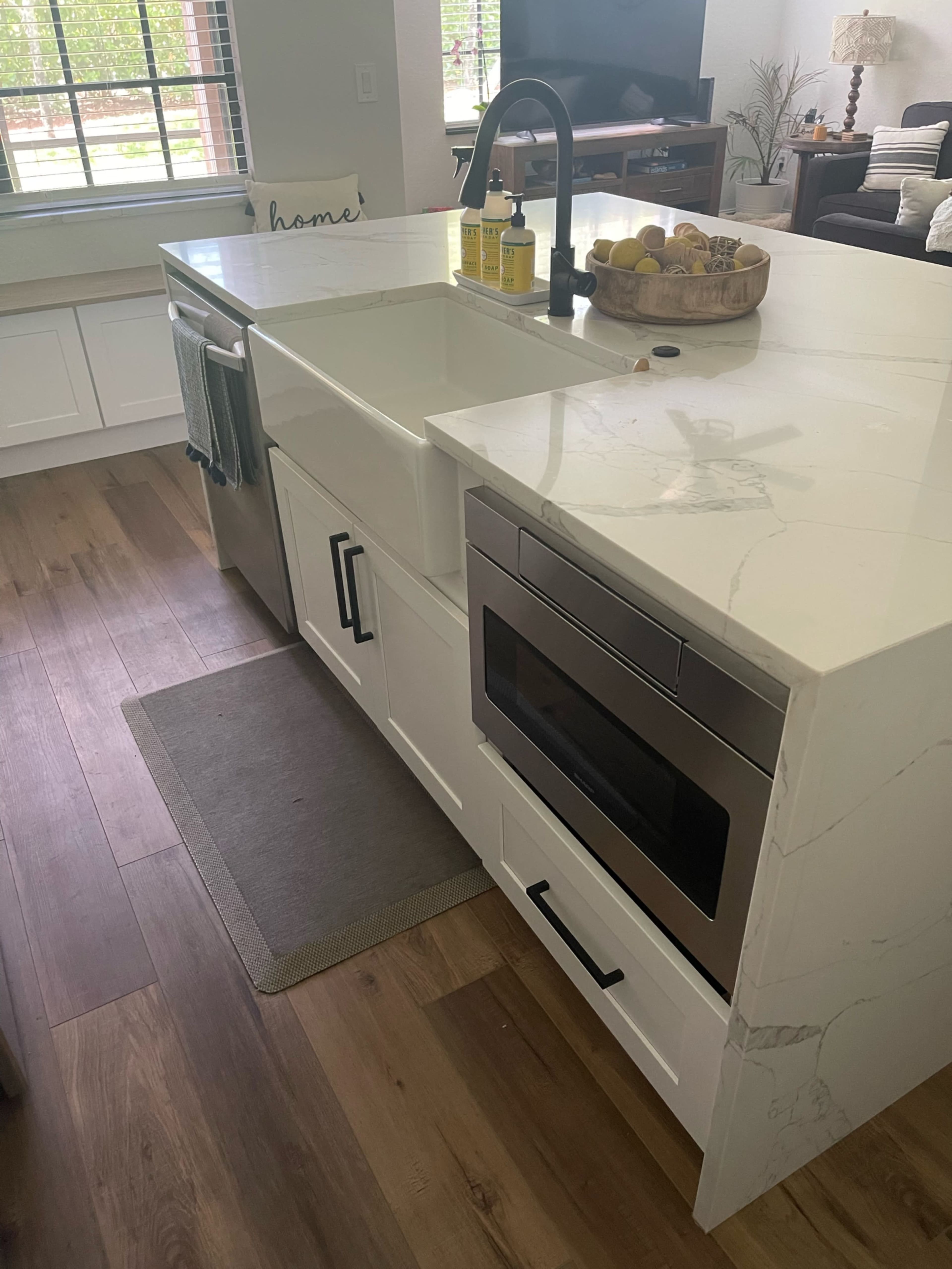 A modern kitchen with a white countertop, a sink, a built-in microwave, and a bowl of fruit on a decorative tray.